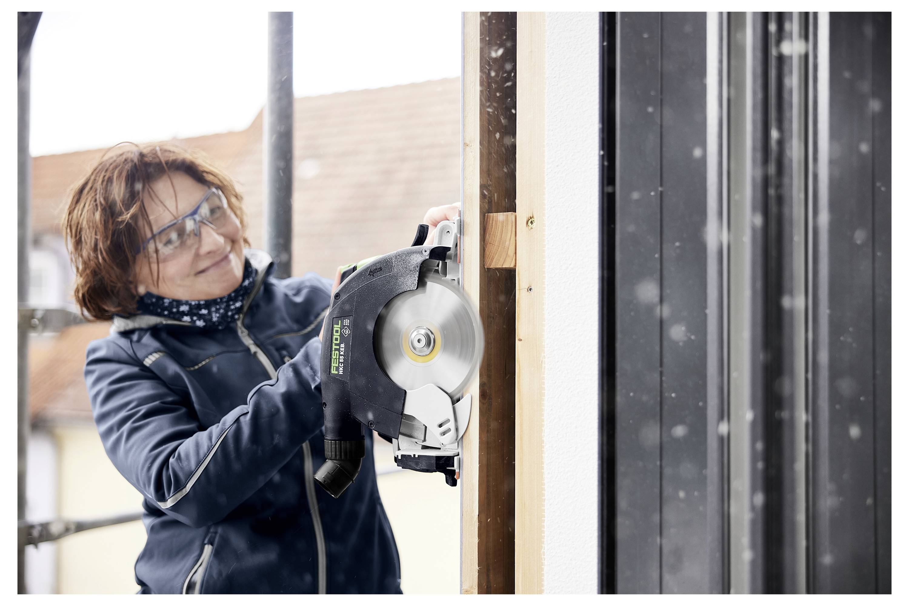 Eine Person, die eine Schutzbrille trägt, benutzt eine Kreissäge an einer Holzkonstruktion und arbeitet an einem verschneiten Tag in einer Baustelle.