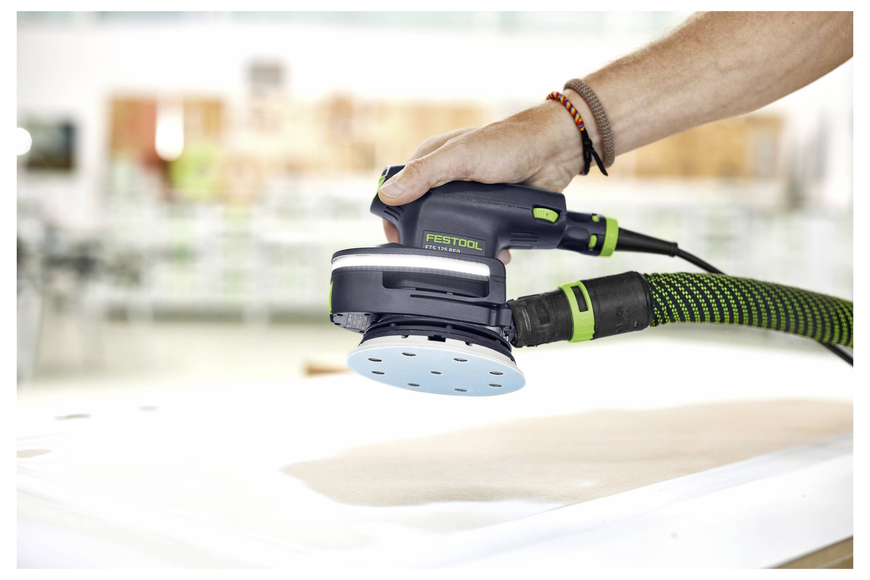 Eine Hand, die einen Festool-Elektroschleiffer hält und auf einer flachen Holzoberfläche arbeitet, mit einem angeschlossenen Schlauch zur Staubabsaugung.