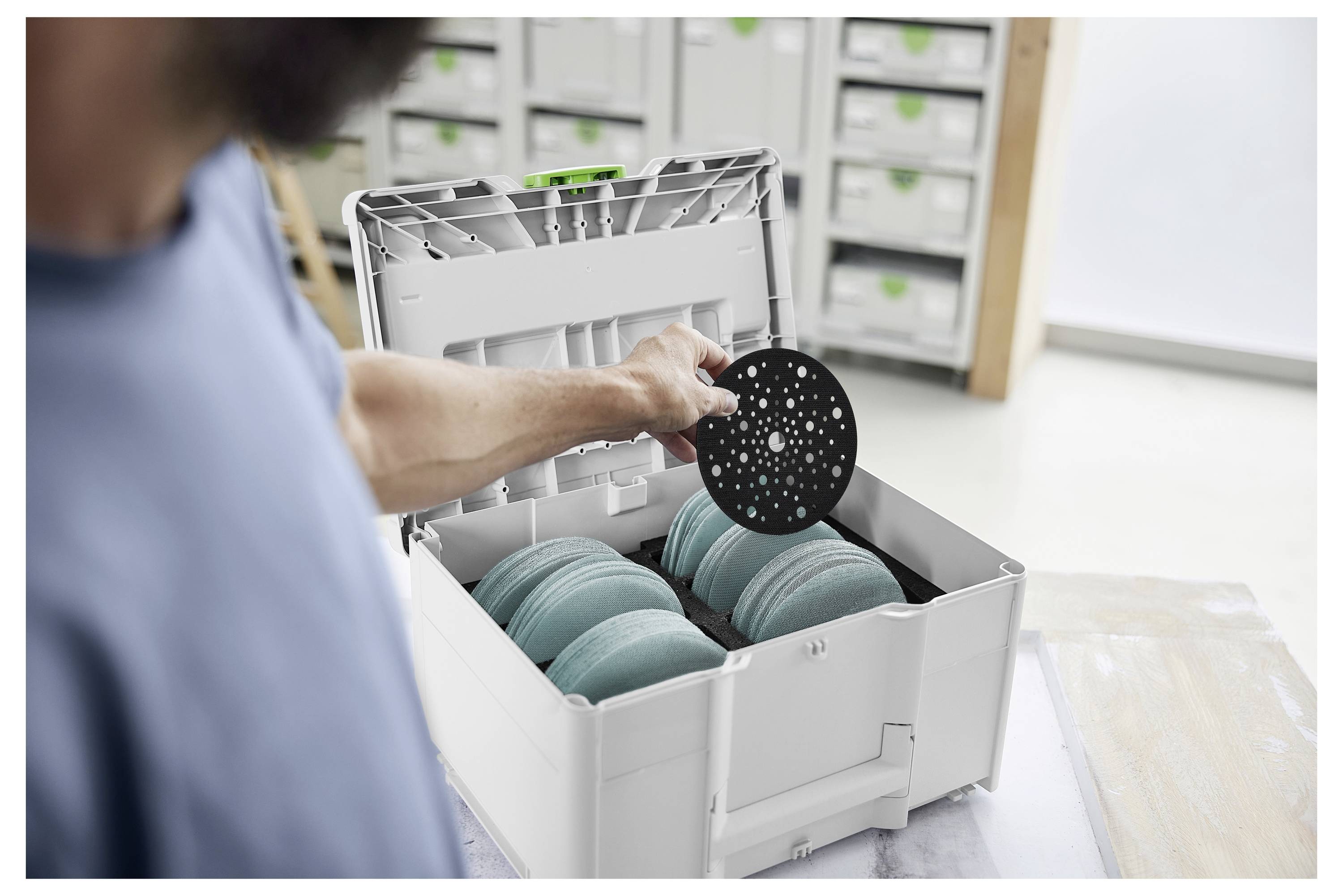 Eine Person legt eine schwarze Kreisscheibe in eine offene Lagerkiste, die mit blaugrünen Scheiben gefüllt ist, in einer gut organisierten Werkstattumgebung.
