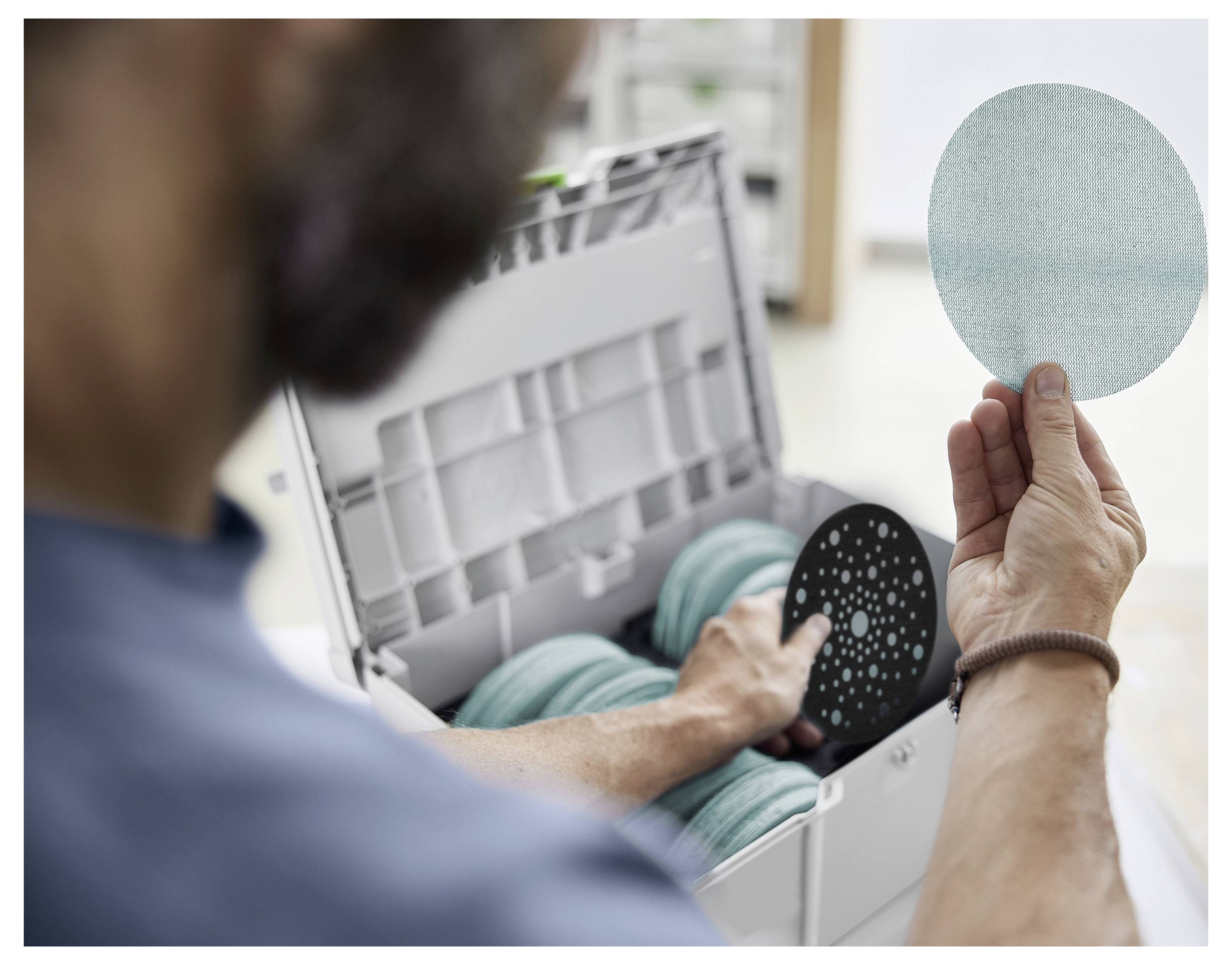 Eine Person hält eine runde, hellblaue Schleifscheibe im Fokus, während weitere Scheiben in einem Behälter gelagert sind. Die Szene deutet auf Vorbereitungen für Schleifarbeiten hin.