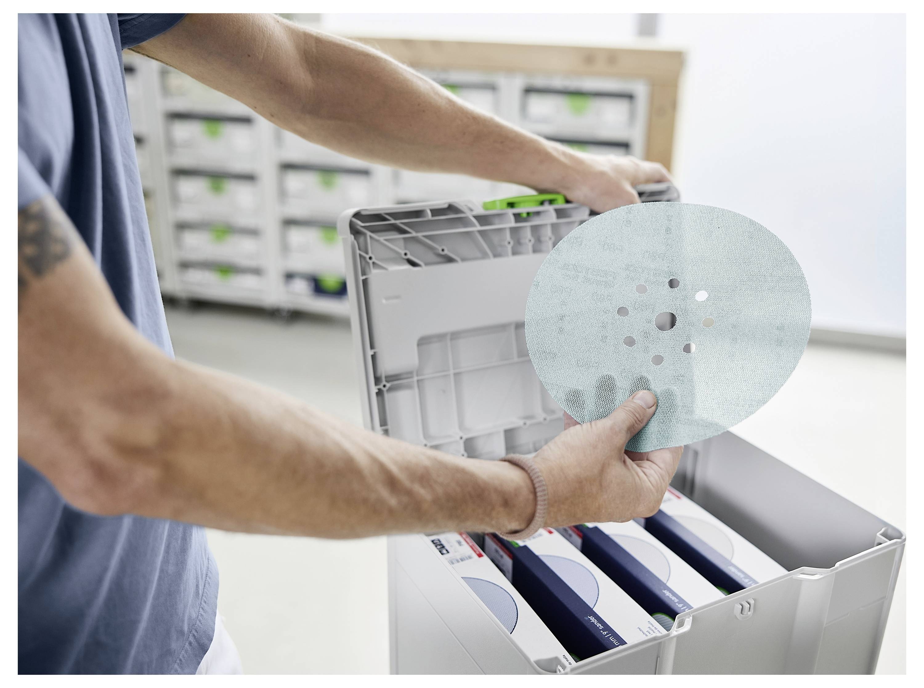 Ein Mann, der eine Schutzbrille trägt, verwendet ein Elektrowerkzeug zum Abschleifen einer Decke und sorgt dabei für eine glatte Oberfläche. Er hält den Staubsaugerschlauch, der mit dem Werkzeug verbunden ist.