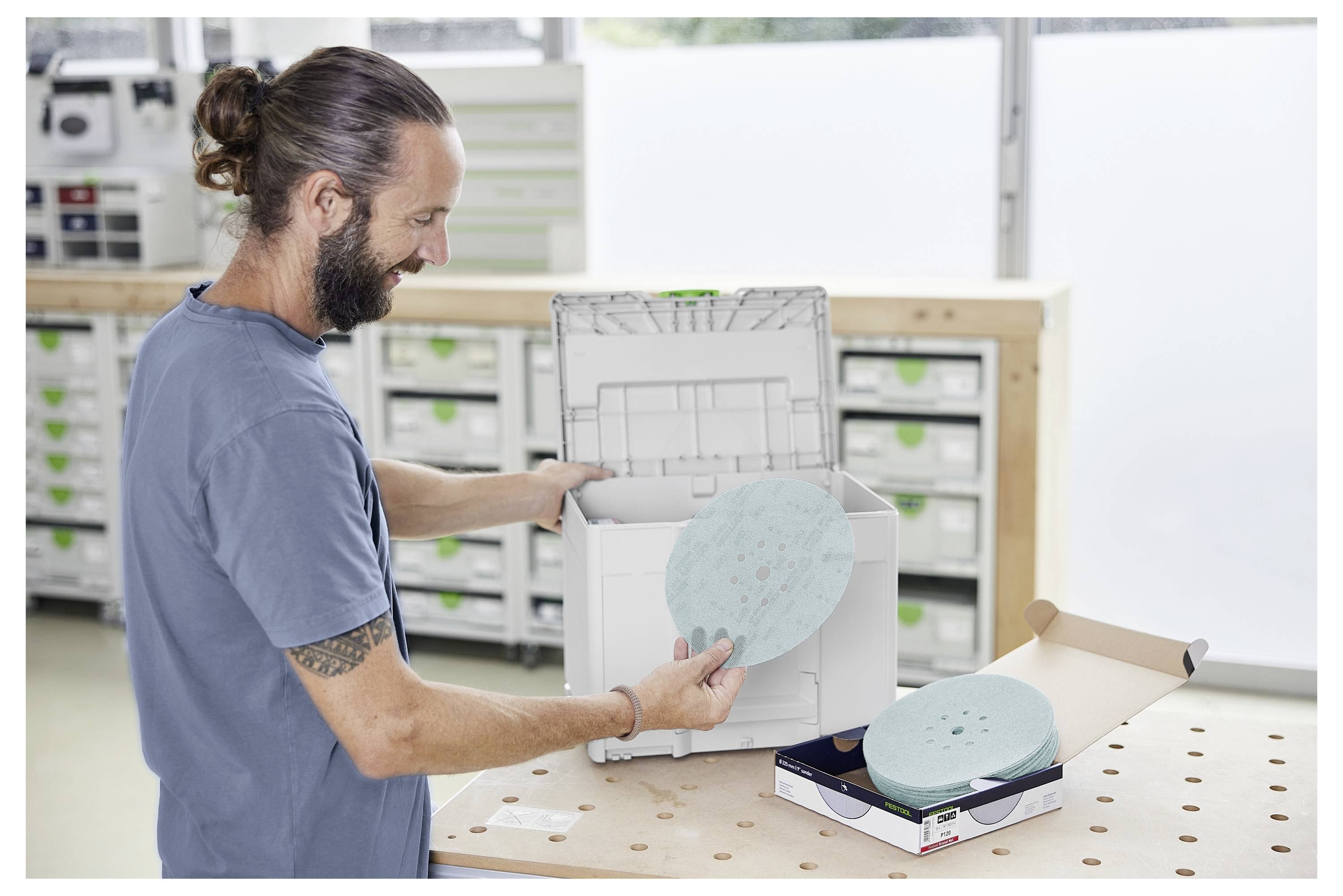 Eine Person mit Tätowierungen bearbeitet eine Holzoberfläche mit einem elektrischen Schleifsystem. Der Fokus liegt auf dem Werkzeug und zeigt dessen Details und Anwendung.