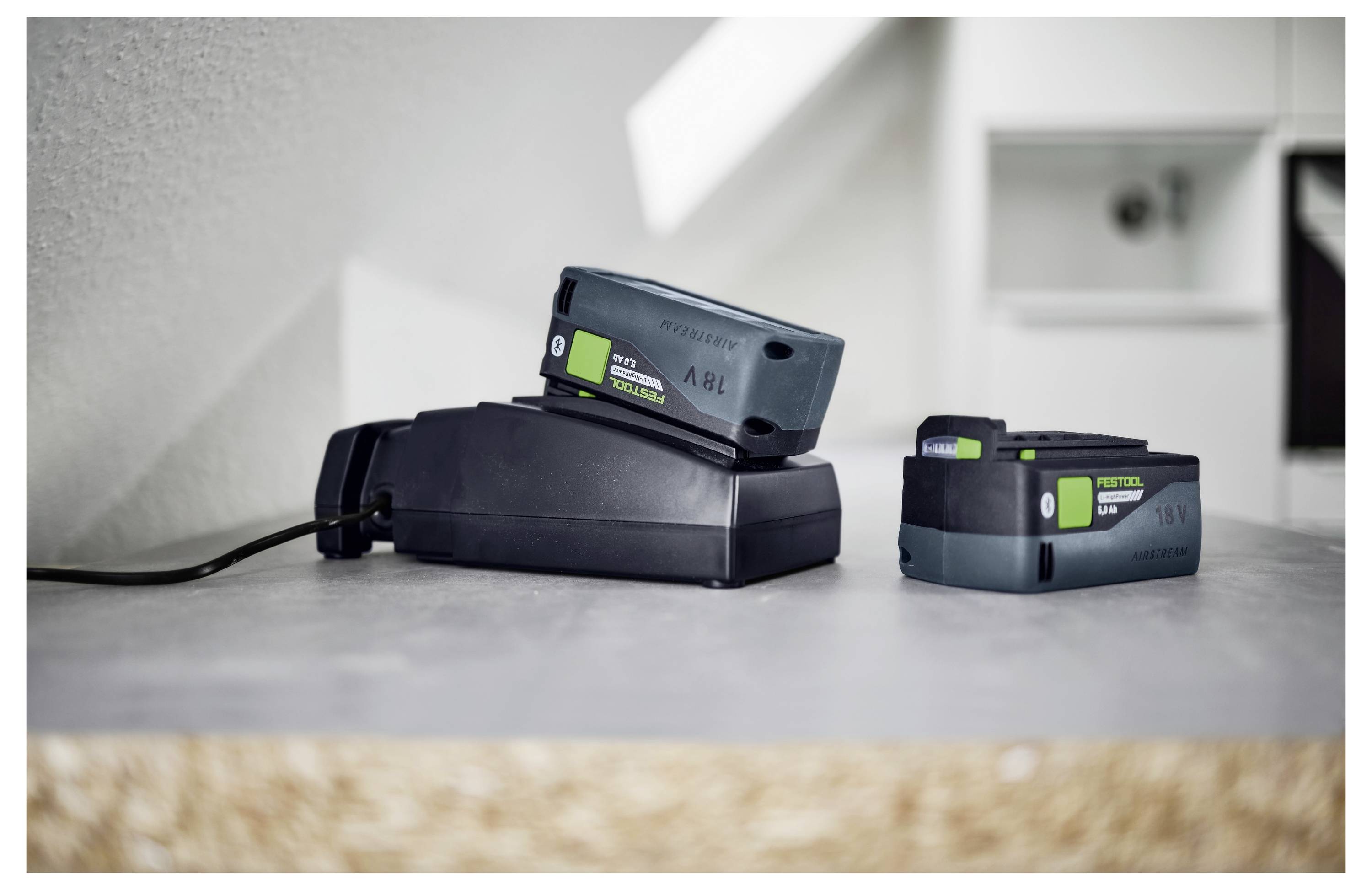 Zwei grün-schwarze Elektrowerkzeugakkus, einer wird geladen und einer steht auf Standby, auf einer grauen Arbeitsplatte in einer Werkstattsituation.