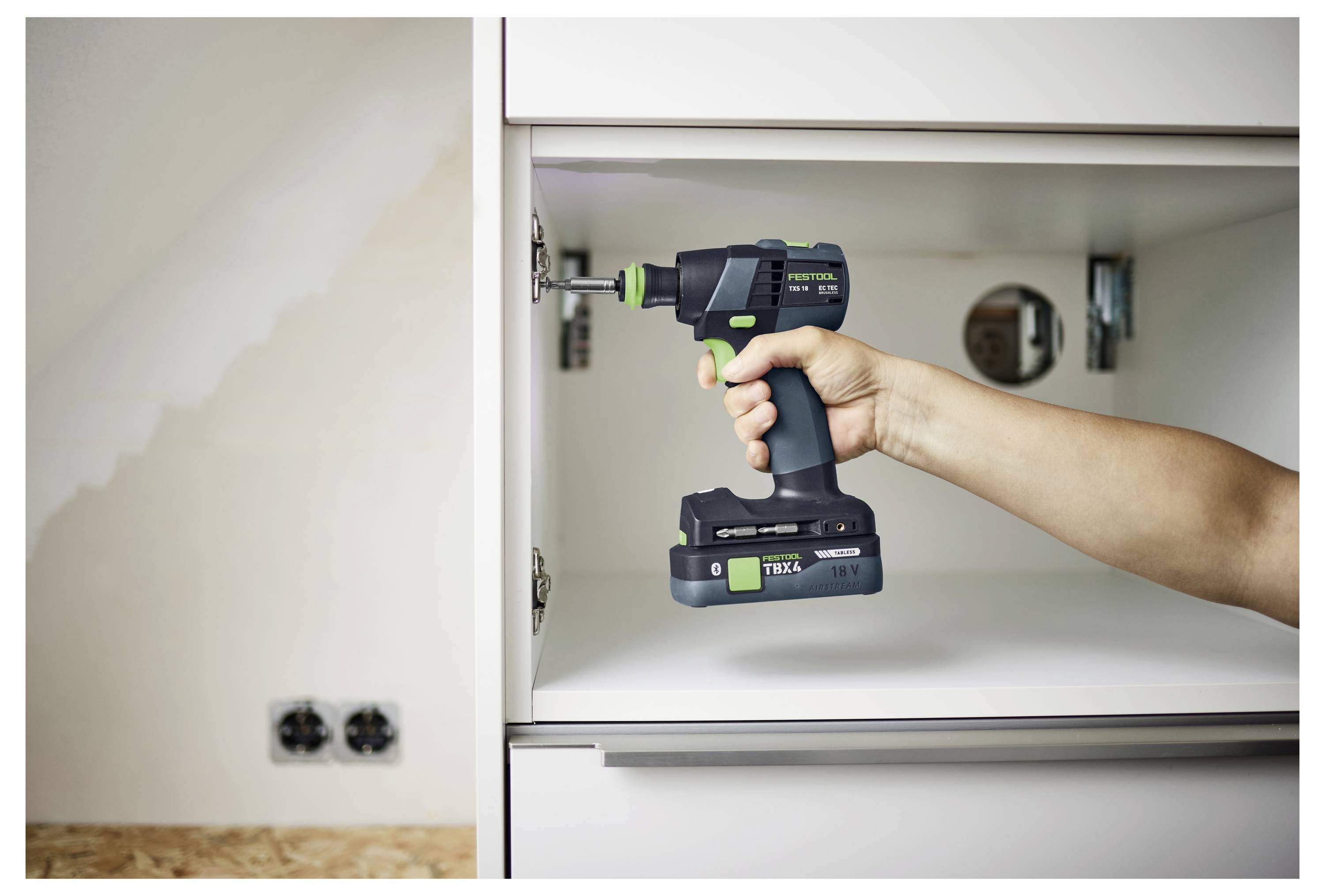 Eine Person benutzt eine Elektrobohrmaschine, um eine Schraube in einen Schrank in einem teilweise fertiggestellten Raum mit sichtbaren Steckdosen und Trockenbau einzufügen.