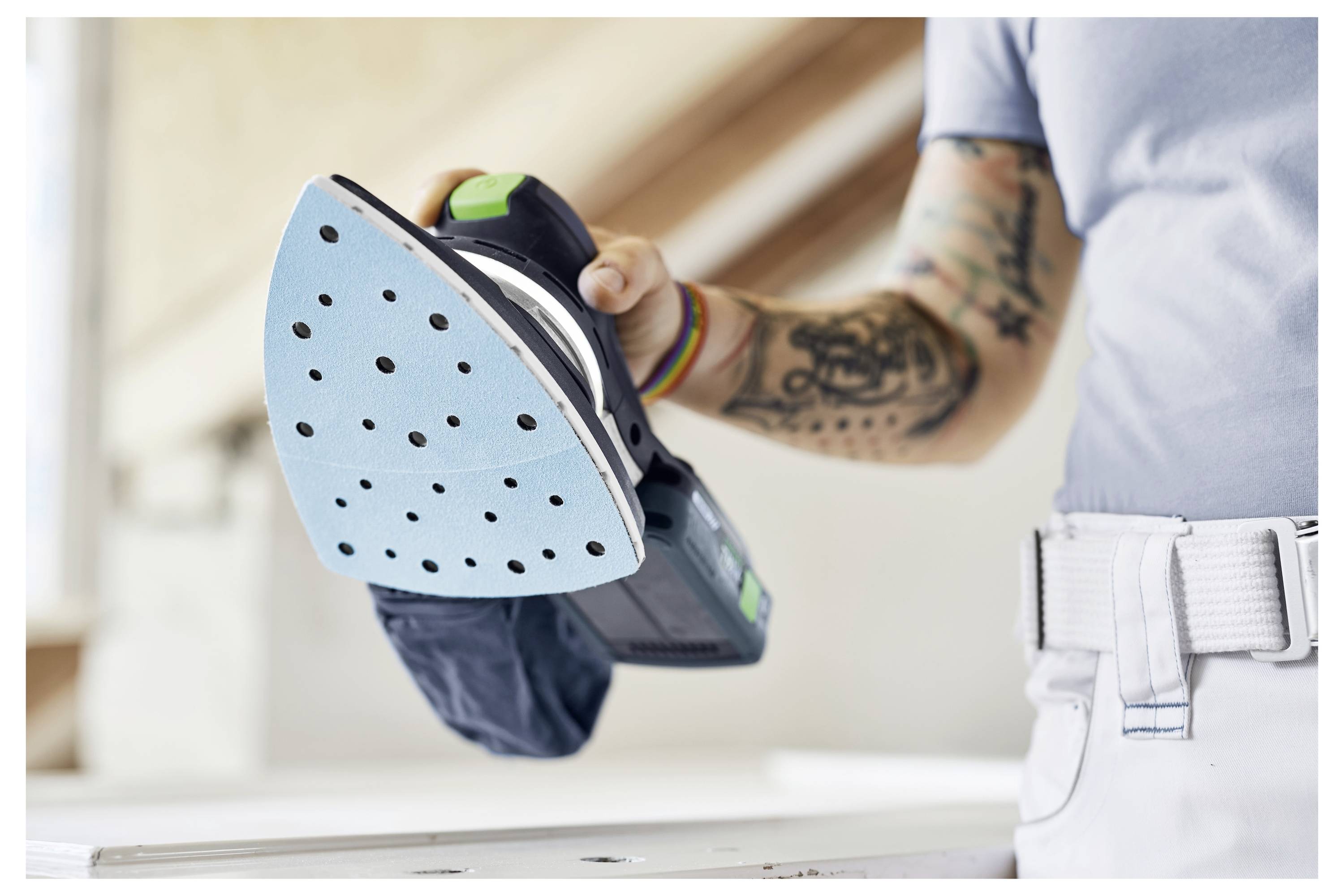 Eine Person in einem hellblauen Hemd hält einen elektrischen Schleifer mit einem blauen Schleifpad. Der Fokus liegt auf dem Werkzeug und ihrem tätowierten Arm.