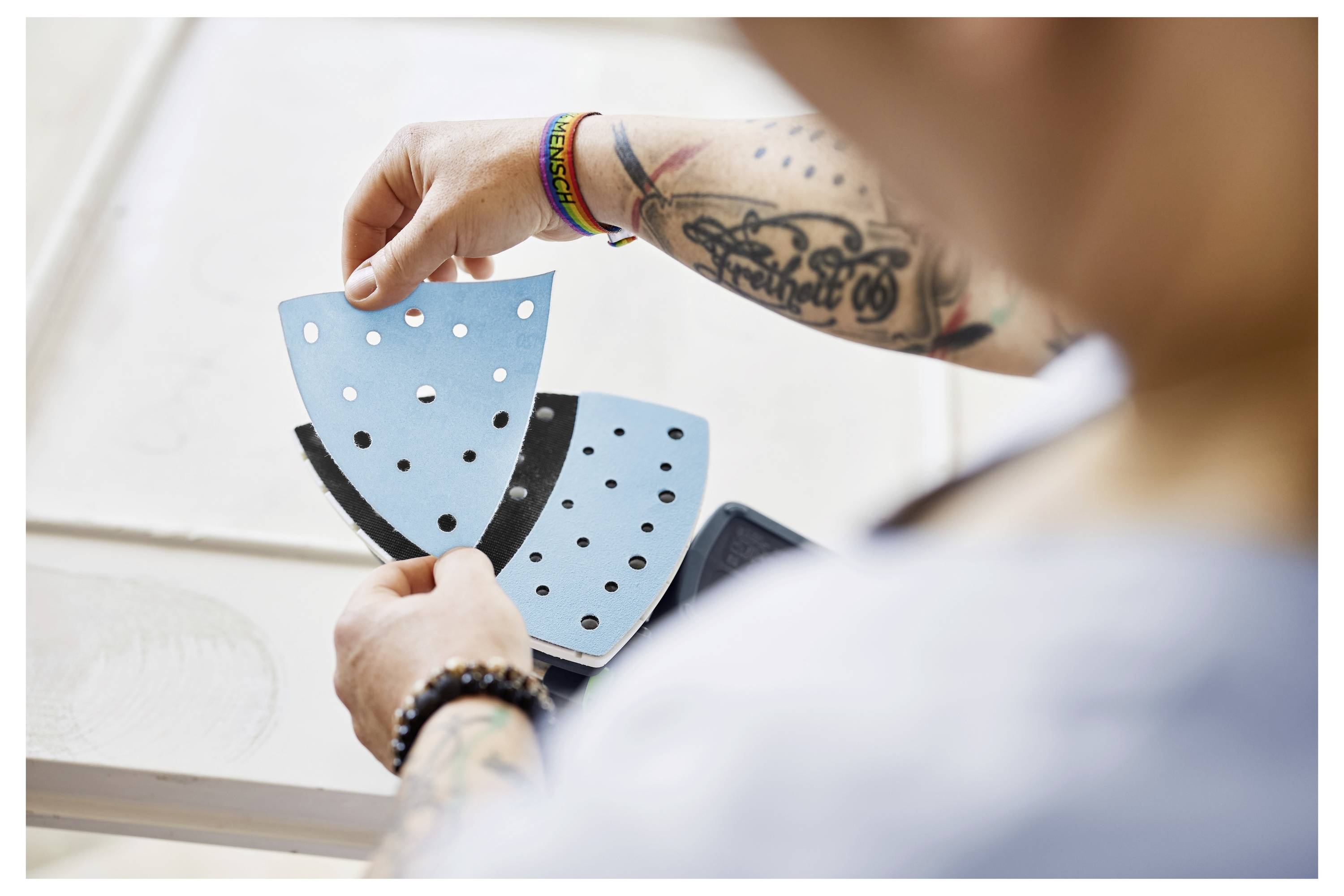 Person, die blaues und schwarzes perforiertes Schleifpapier hält, mit Tattoos und mit einem mehrfarbigen Armband.