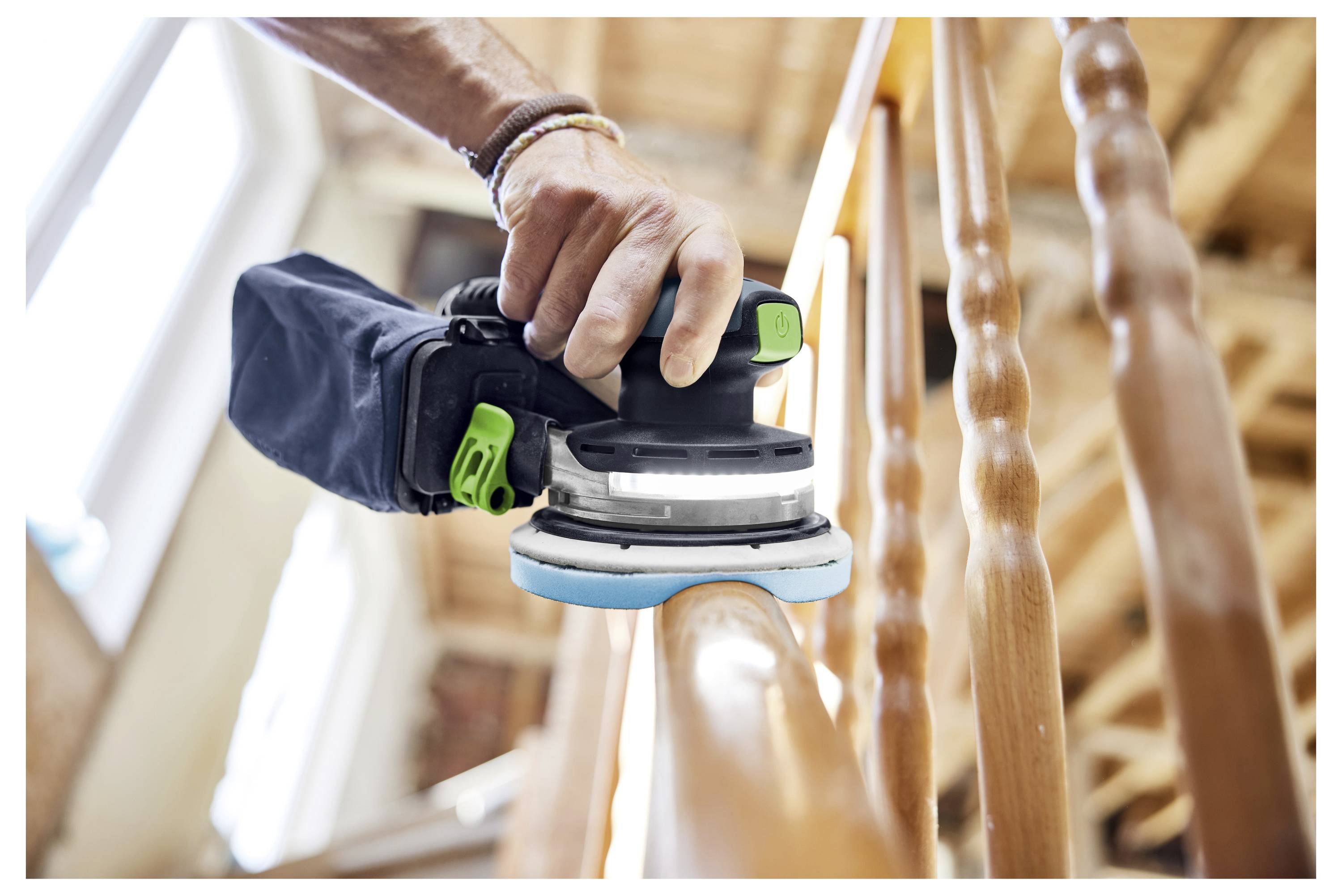 Eine Person benutzt einen tragbaren Elektroschleiffer, um ein Holzgeländer einer Treppe in einem Raum mit Holzbalken zu glätten.