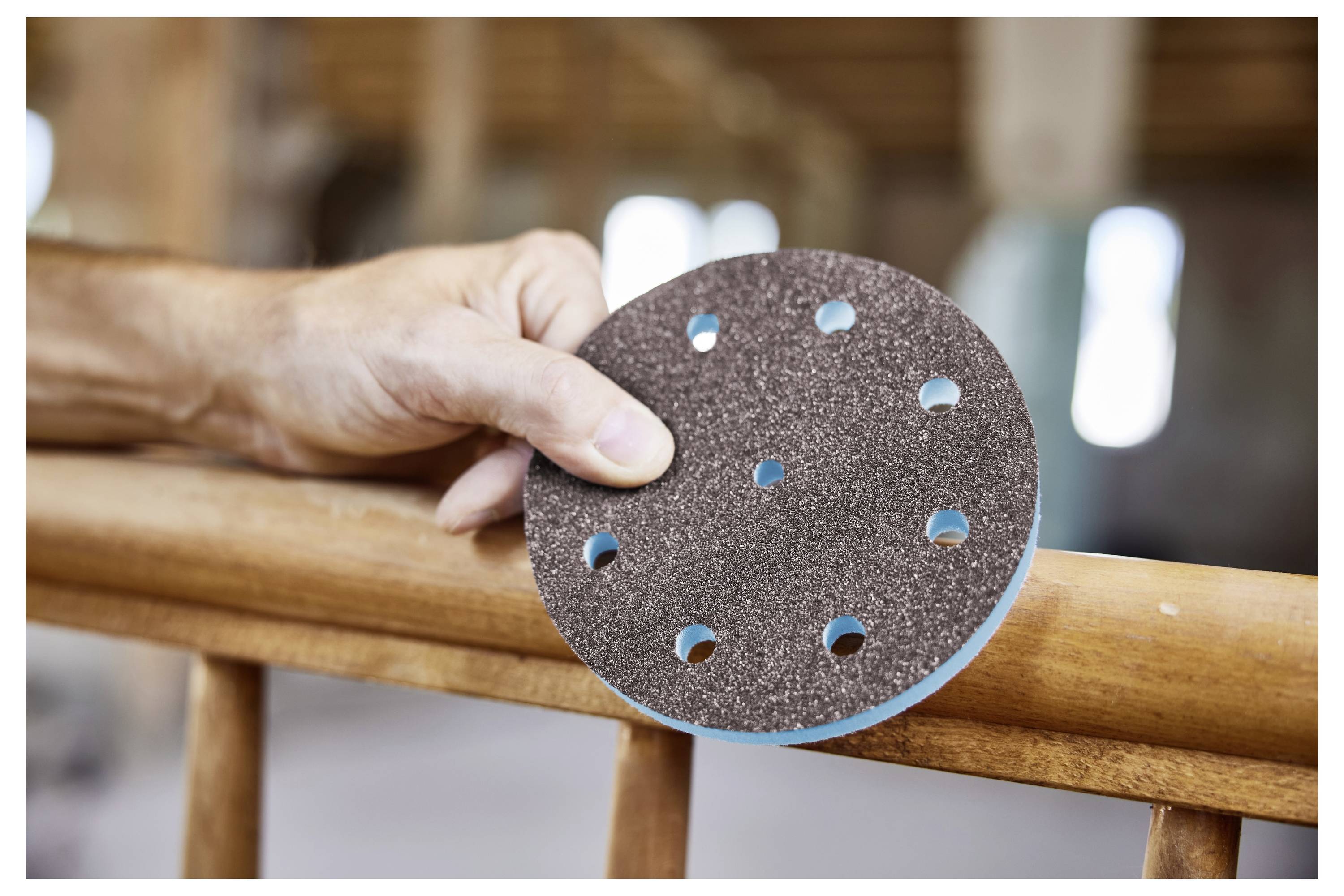 Eine Hand hält ein rundes Schleifpad mit Löchern und positioniert es gegen ein hölzernes Geländer, was die Vorbereitung für Schleifarbeiten anzeigt.
