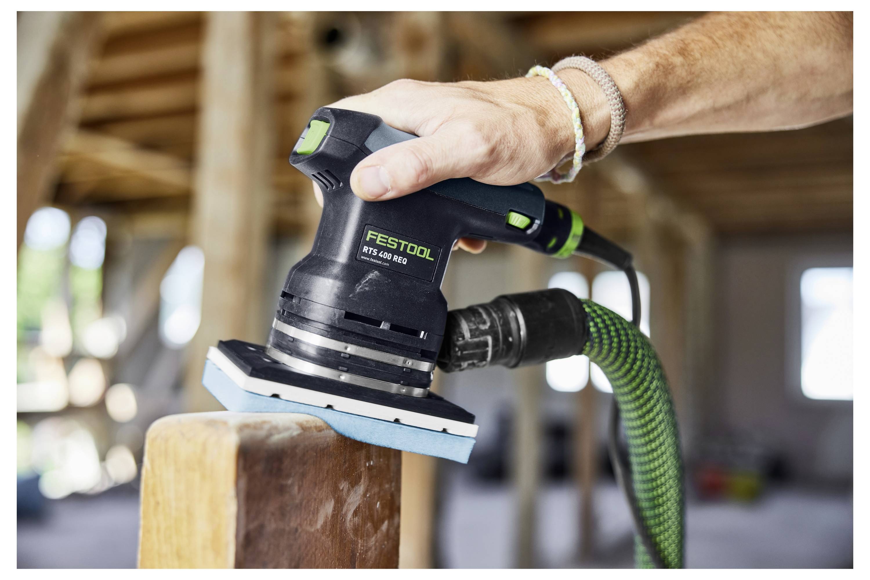Eine Hand benutzt einen Festool-Elektroschleiffer auf einem Holzbalken in einer Baustelle. Sonnenlicht fällt durch Fenster im Hintergrund herein.