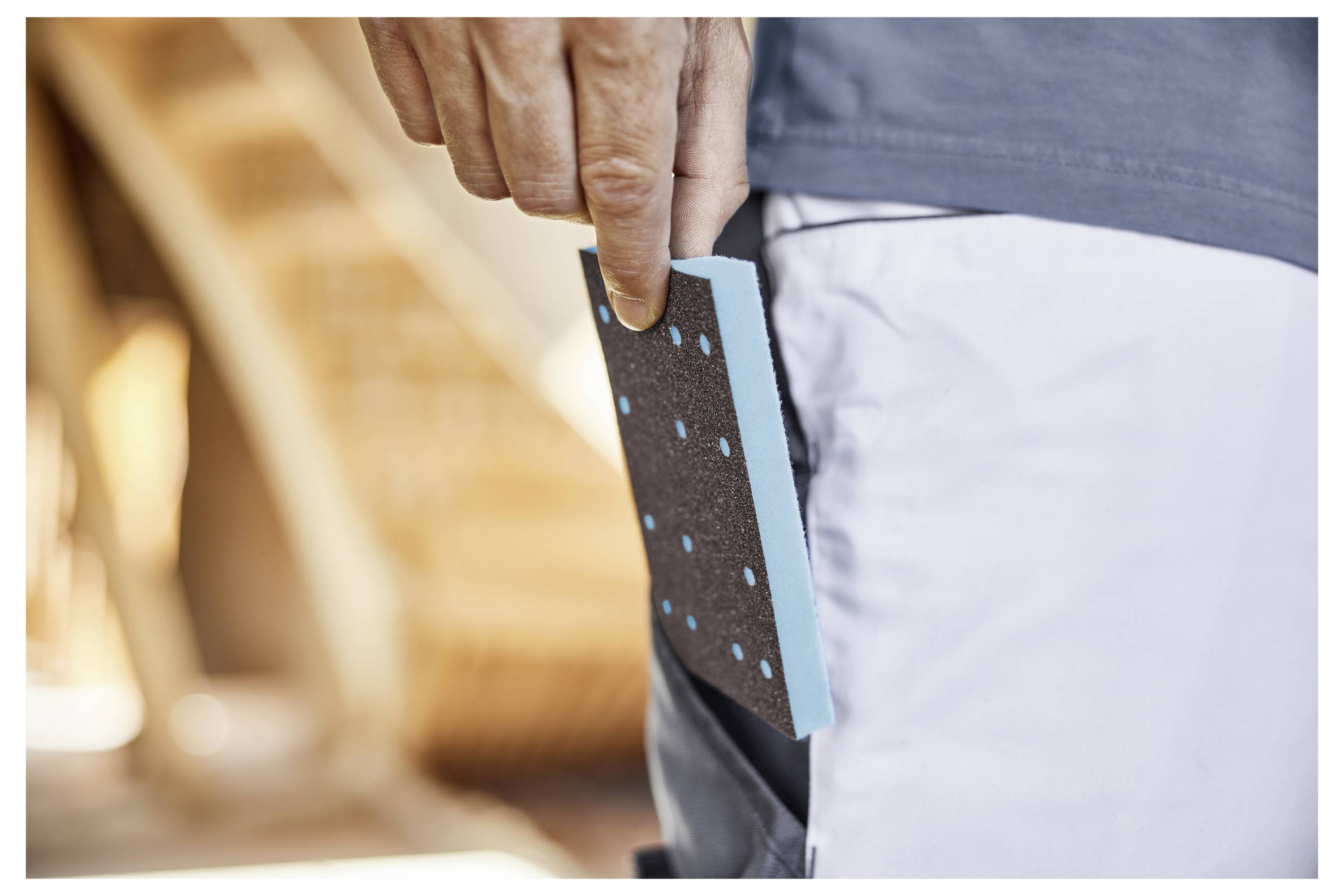 Eine Hand benutzt einen elektrischen Elektroschleiffer auf einer Holzoberfläche und glättet das Holz. Das Werkzeug ist schwarz mit grünen Akzenten, und ein Staubsammelbeutel ist angebracht.