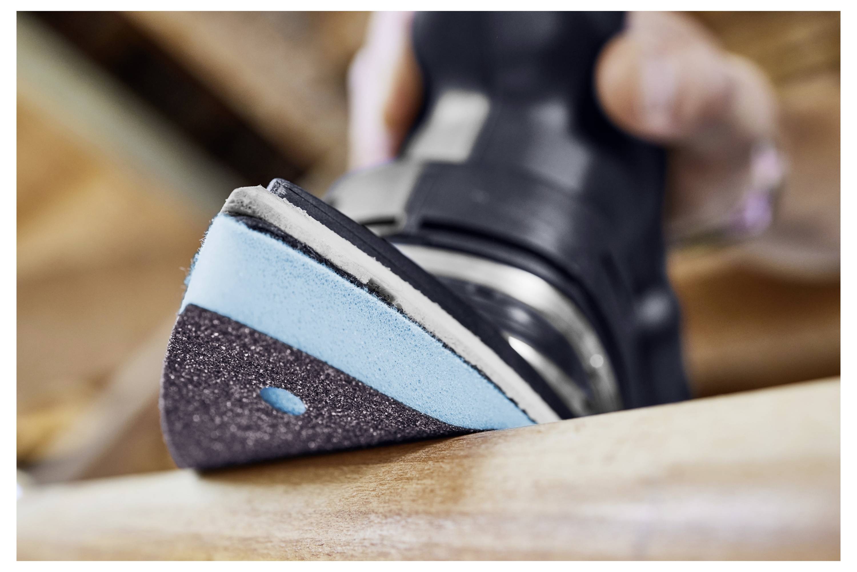 Eine Nahaufnahme zeigt eine Hand, die einen Elektroschleiffer hält und die Oberfläche einer Holzplanke glättet, wobei das blaue und schwarze Schleifpad sichtbar wird.