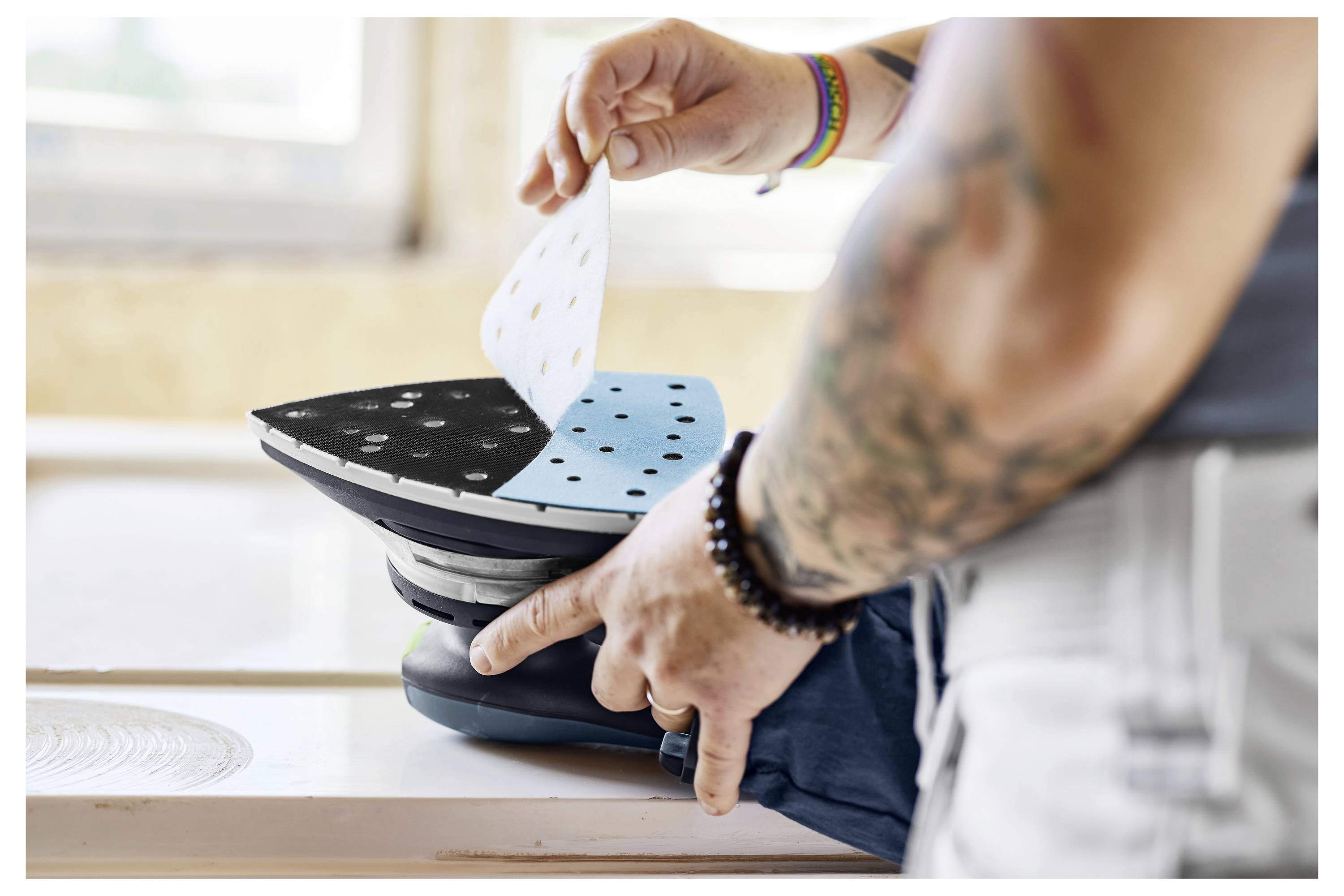 Eine Person bringt Schleifpapier auf einem Schleifwerkzeug an und bereitet es zur Verwendung vor. Helle Armbänder und Tätowierungen sind am Arm der Person sichtbar.