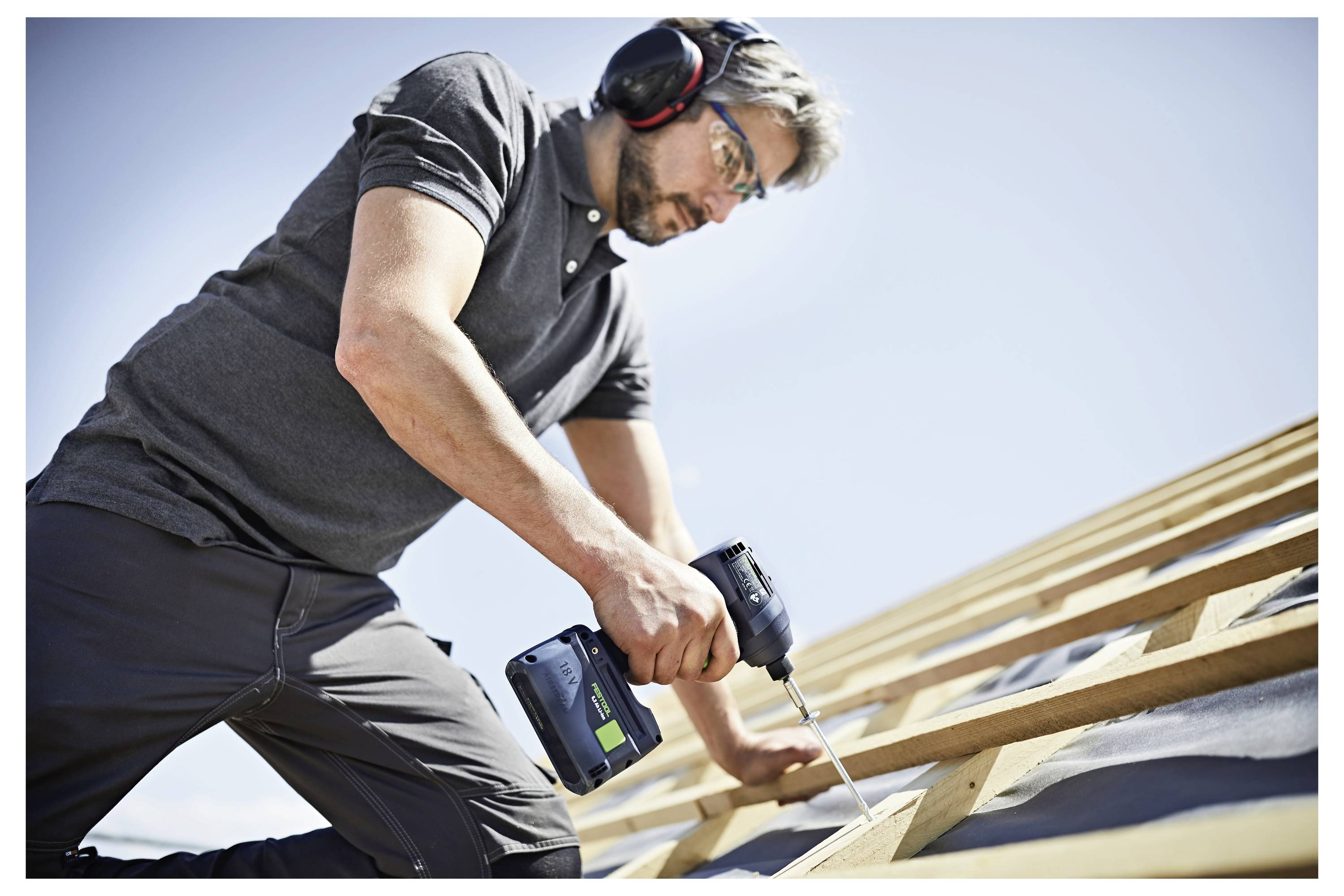 Eine Person in Schutzausrüstung verwendet eine Elektrobohrmaschine, um Balken auf einem Dach unter einem klaren blauen Himmel zu befestigen.