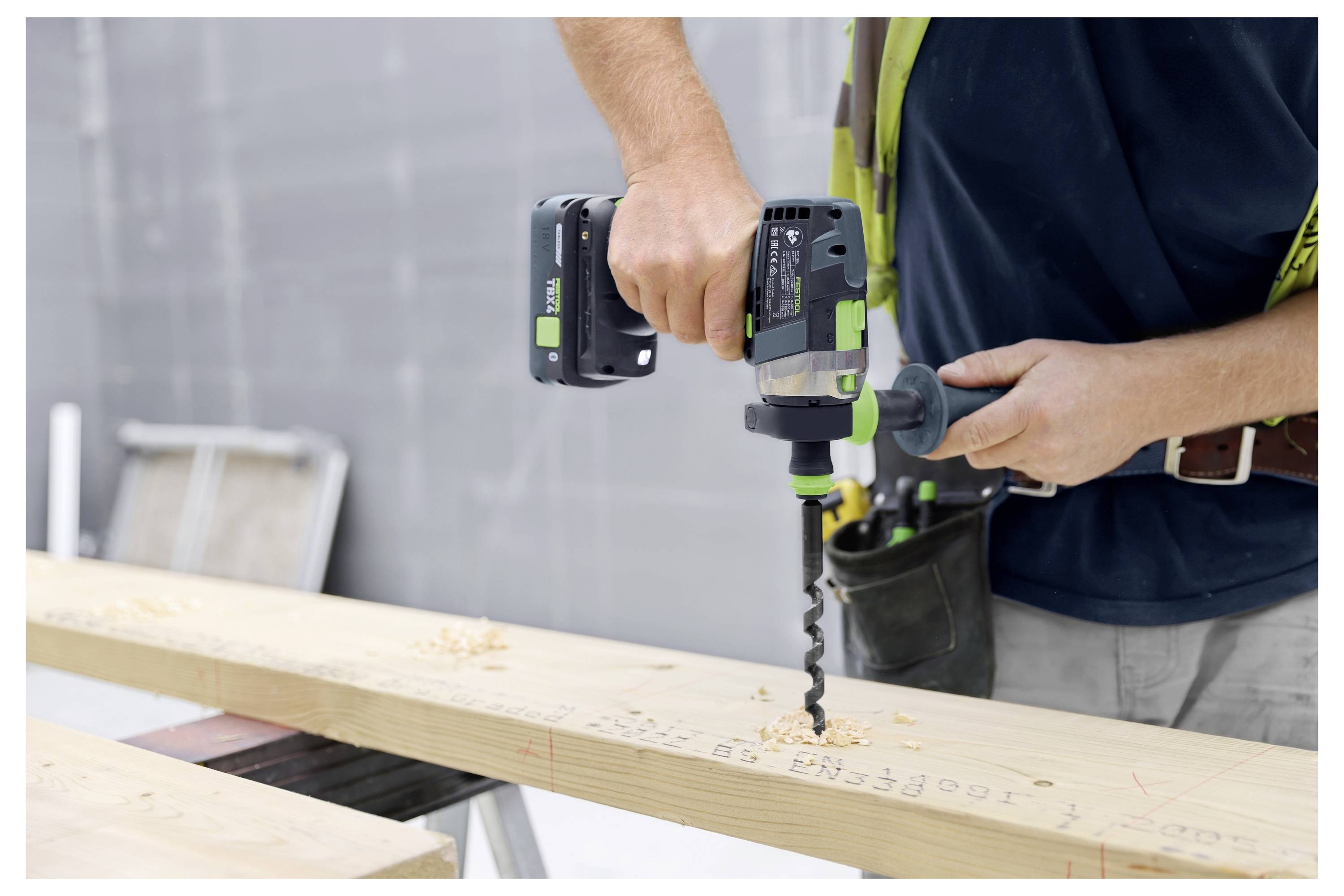 Eine Person verwendet einen grün-schwarzen Elektrobohrer, um in ein Stück Holz zu bohren. Holzspäne sind sichtbar. Die Umgebung sieht wie eine Werkstatt aus.
