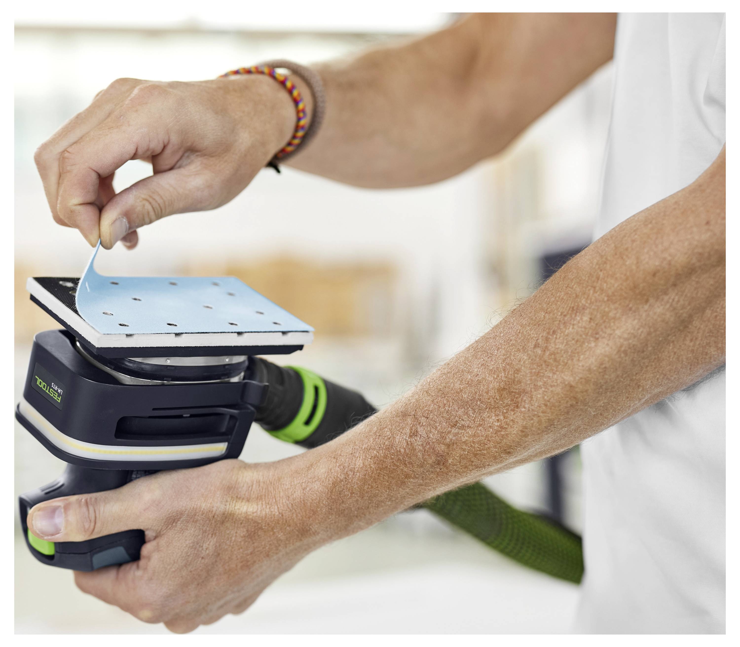 Eine Person hält einen elektrischen Schleiffer und bringt ein Schleifblatt am Werkzeug an. Der Hintergrund ist verschwommen und deutet auf eine Werkstattumgebung hin.