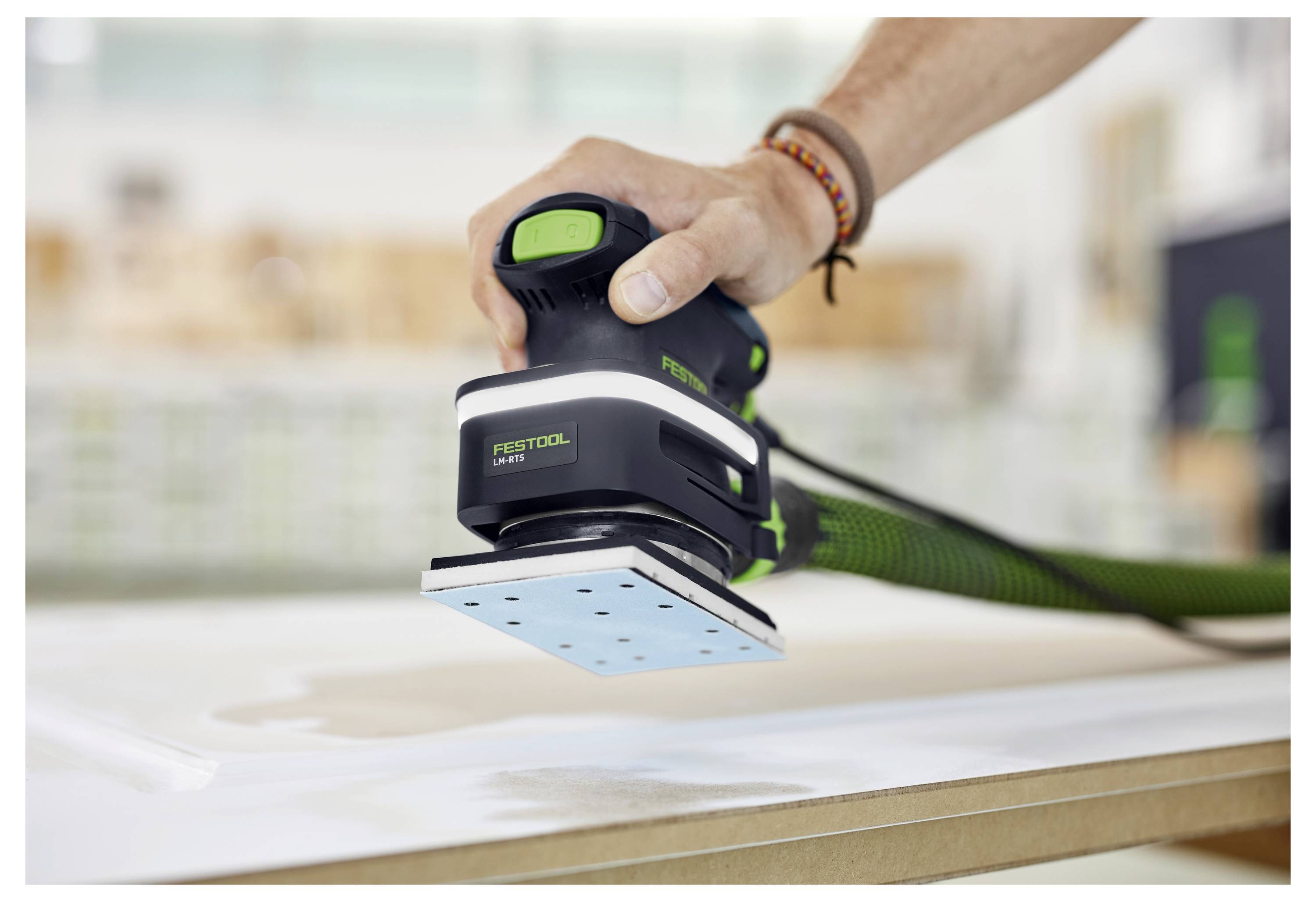 Eine Hand benutzt einen elektrischen Schleifer, um die Oberfläche einer Holzplatte in einer Werkstatt zu glätten.