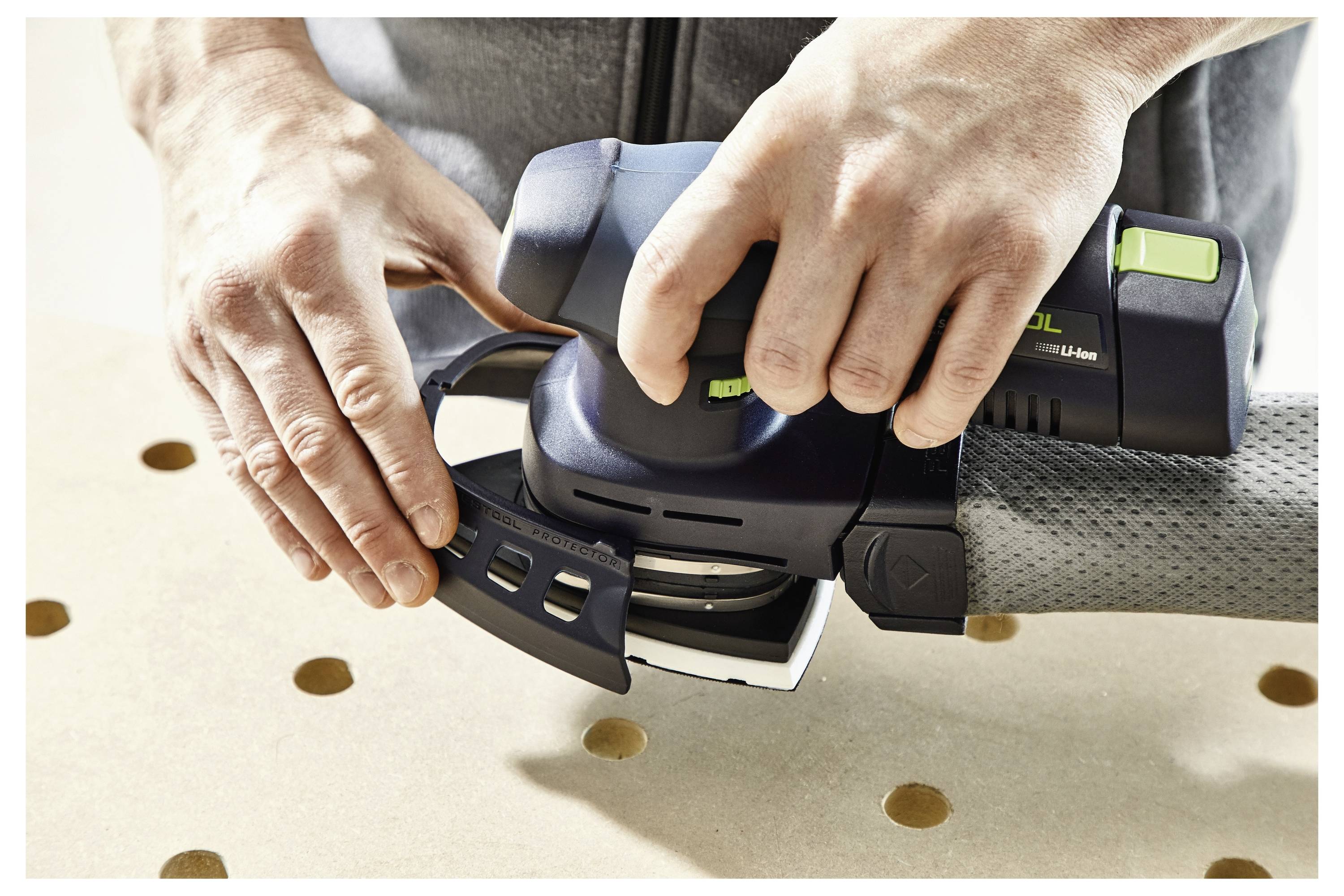 Eine Person benutzt einen tragbaren elektrischen Schleiffer auf einer Holzoberfläche mit Löchern und konzentriert sich darauf, ein abnehmbares Teil des Werkzeugs einzustellen.