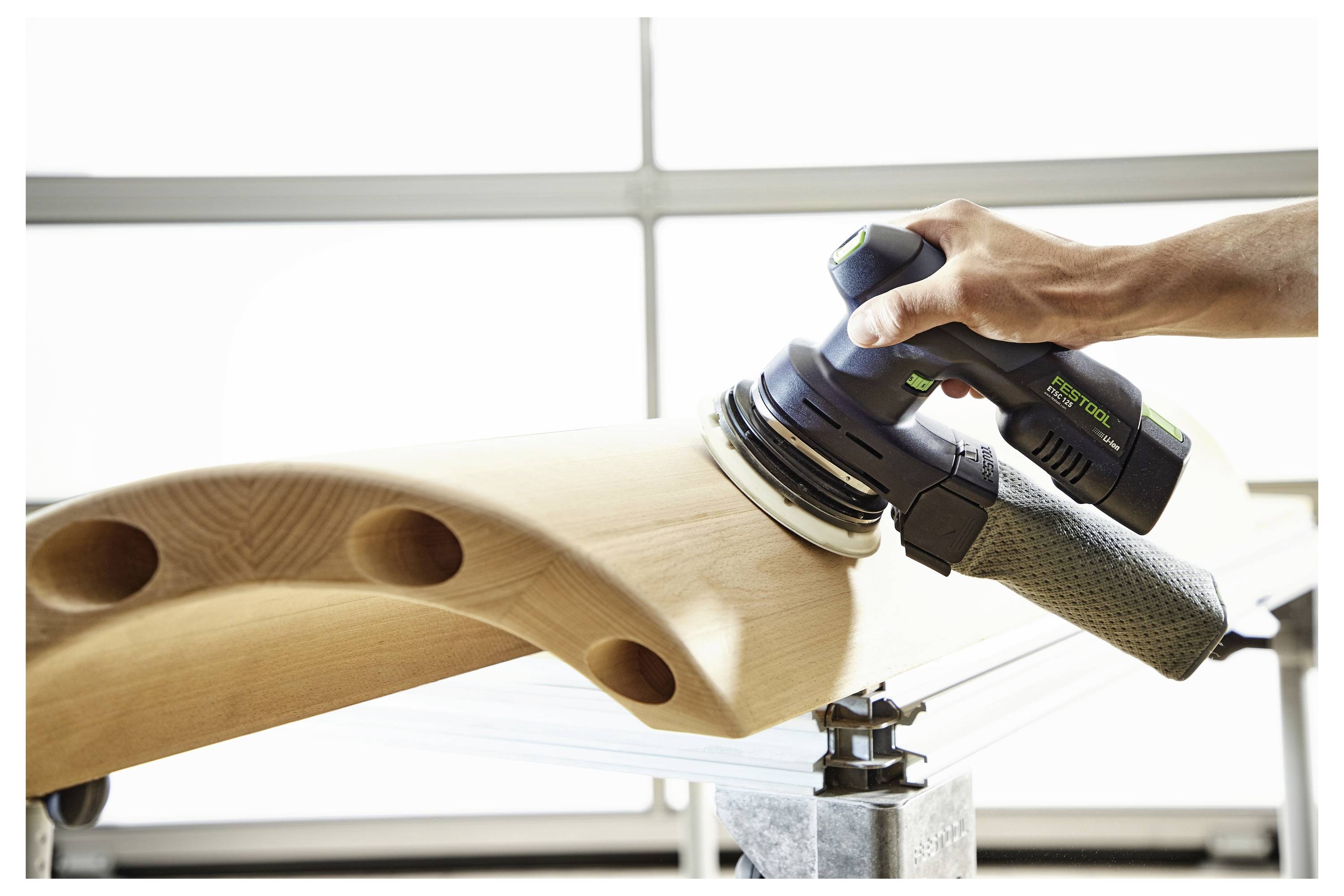 Hand, die einen Schleifklotz hält, teilweise in eine weiße Werkzeugtaschentasche eingeführt, mit unscharfer Holzstruktur im Hintergrund.