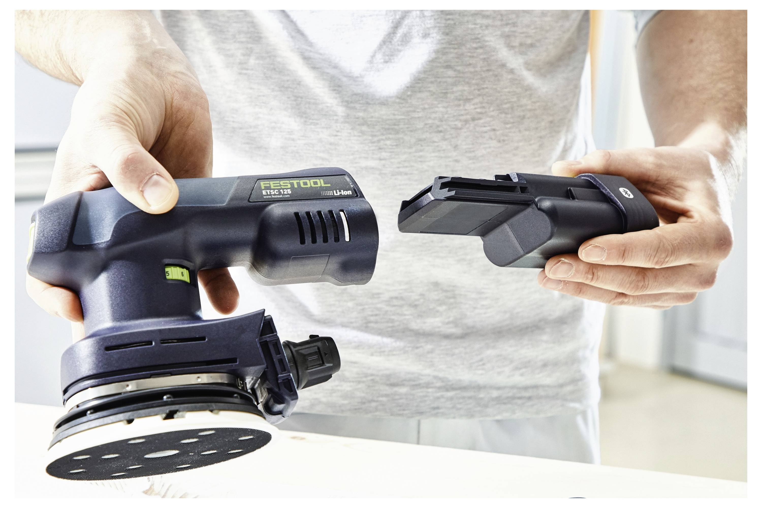 Eine Person, die in einer Hand einen schwarzen tragbaren Elektroschleiffer und in der anderen Hand eine abnehmbare Batterie hält, was darauf hindeutet, dass das Werkzeug mit Batterie betrieben wird.