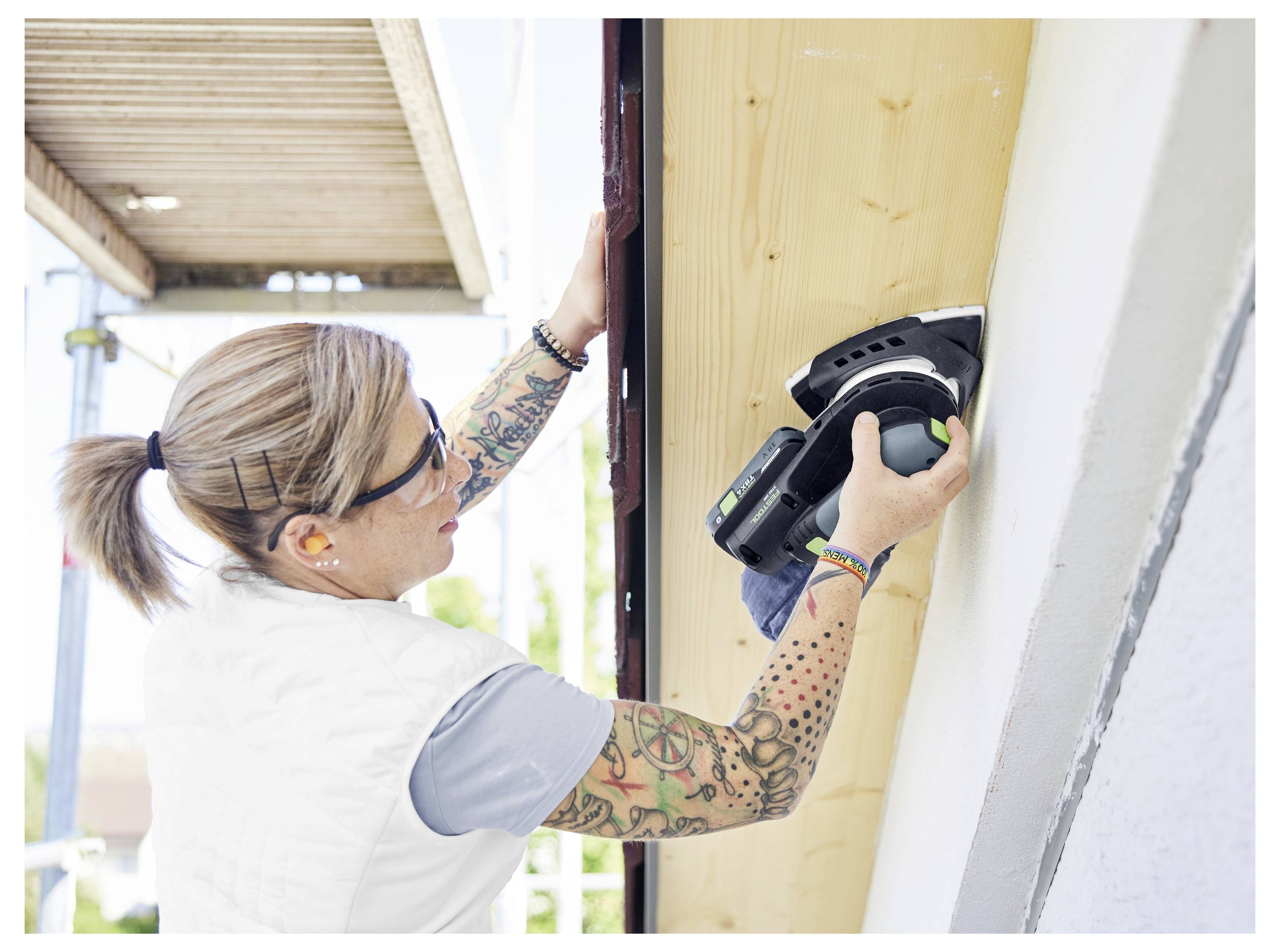 Eine Person, die Schutzbrille und Kopfhörer trägt, verwendet ein Elektrowerkzeug, um eine hölzerne Platte im Freien zu schleifen. Sie hat Tätowierungen auf ihrem linken Arm.