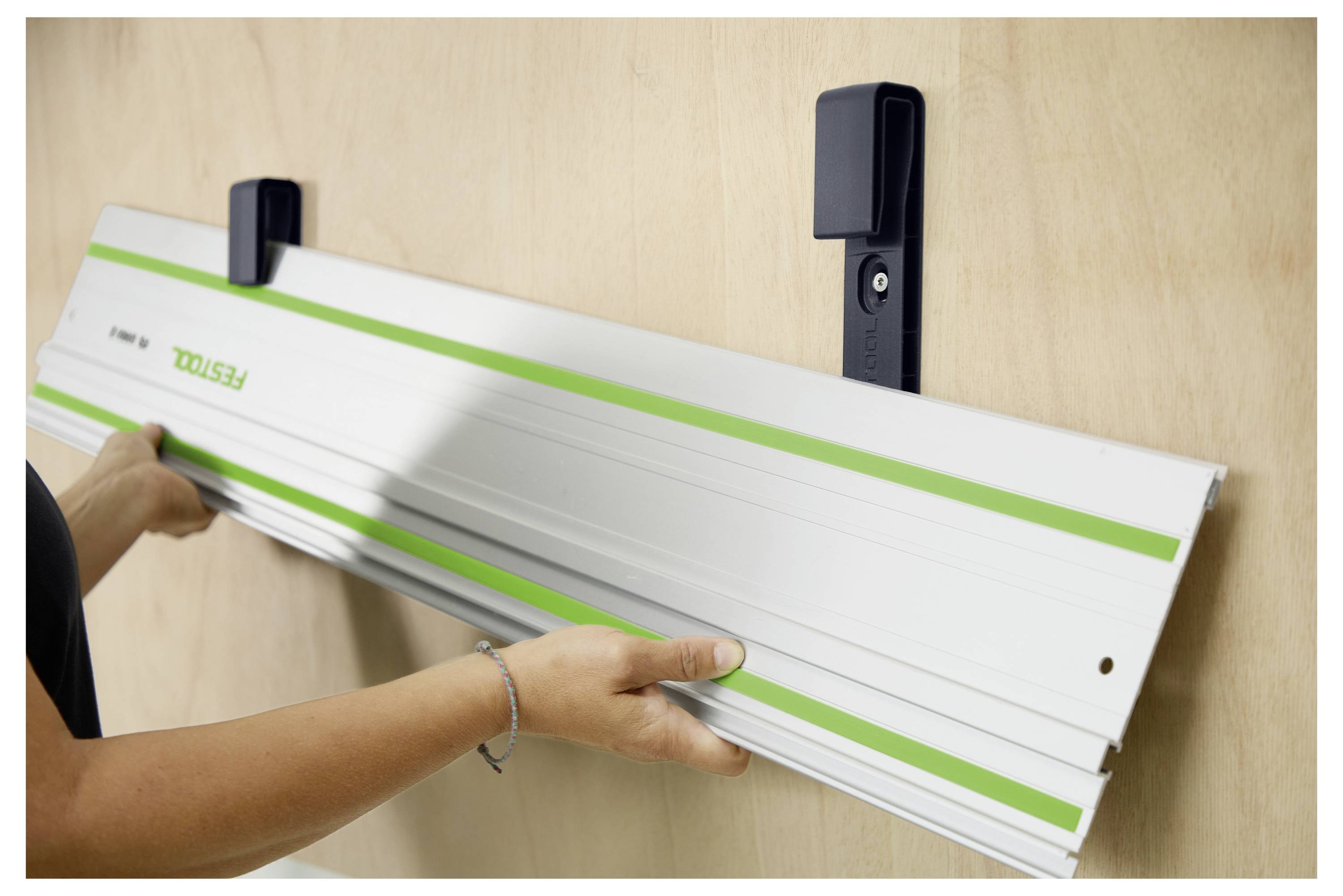 Eine Person befestigt eine lange Metallschiene mit grünen Streifen mit zwei schwarzen Halterungen an einer Wand, möglicherweise für ein Werkzeug- oder Schienensystem.