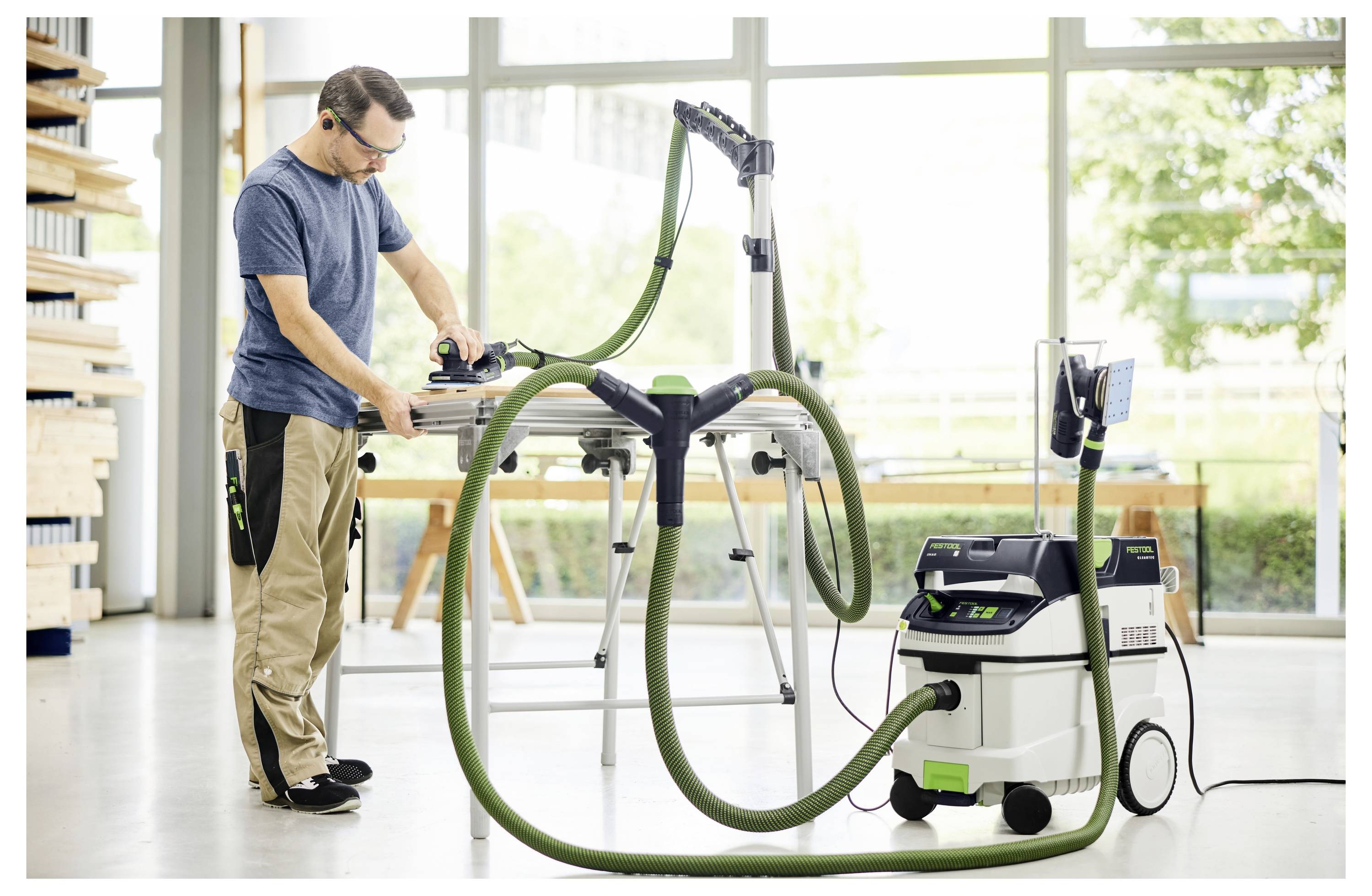 Eine Person mit Schutzbrille schleift einen Holzfensterrahmen in einer Werkstatt, die mit Werkzeugen gefüllt ist und helles natürliches Licht von großen Fenstern erhält.