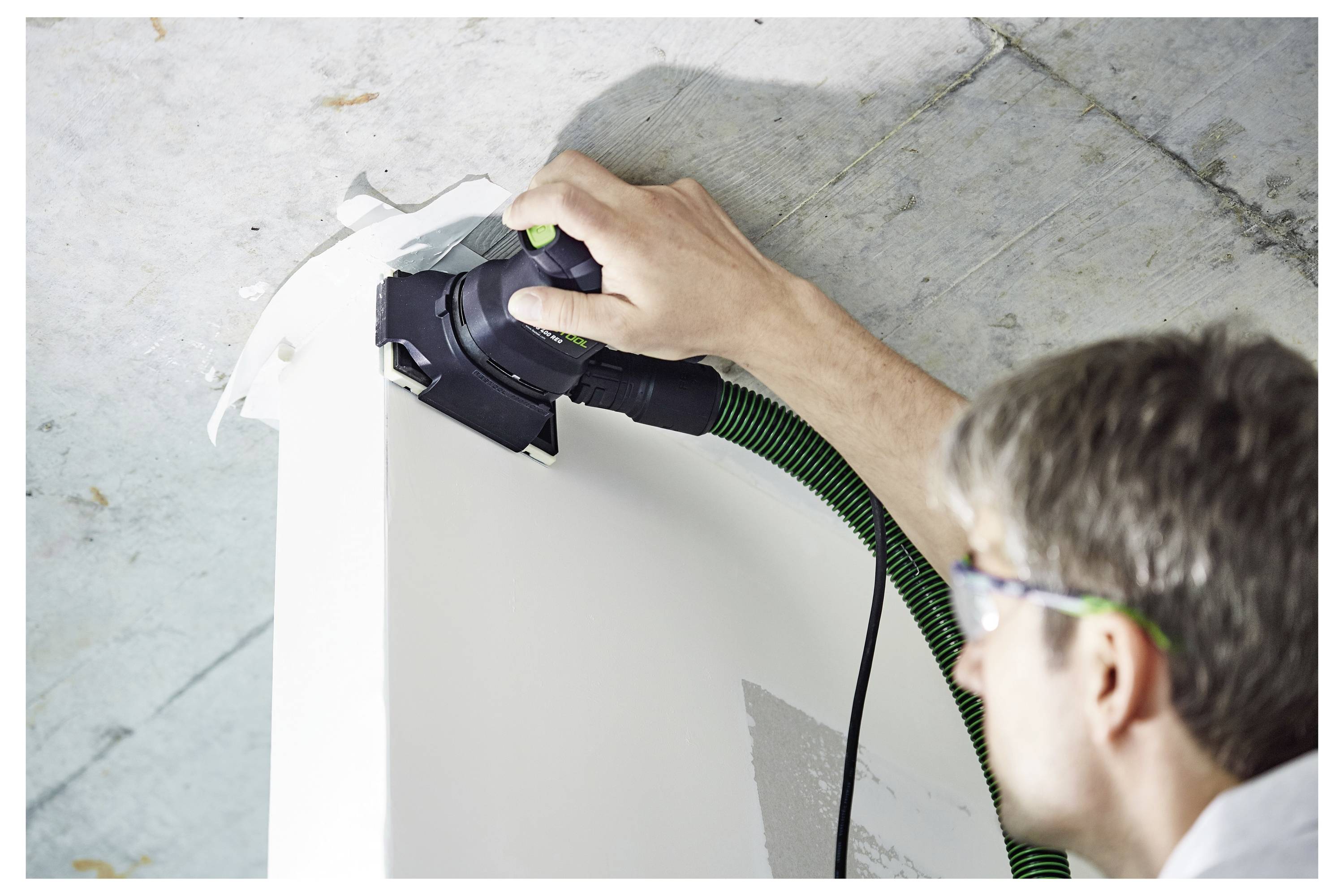 Eine Person mit Schutzbrille benutzt ein Elektrowerkzeug zum Schleifen einer weißen Oberfläche, mit einem grünen Schlauch, der an das Werkzeug angeschlossen ist, unter einer Decke.