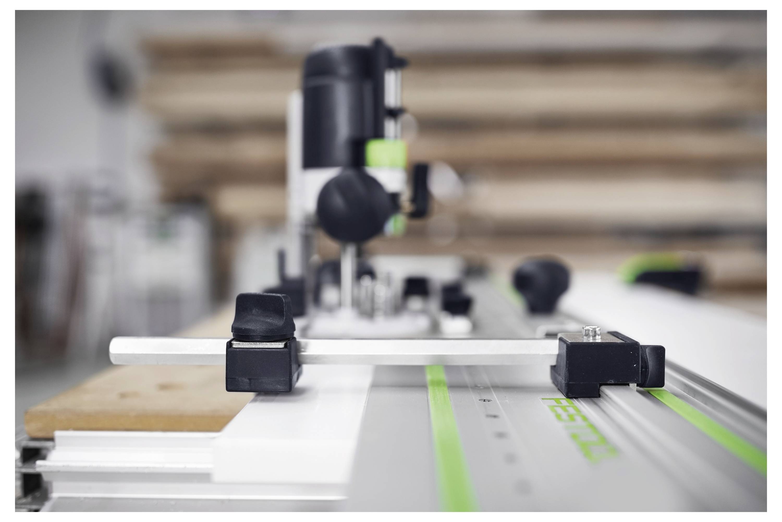 Nahaufnahme einer Oberfräse, die auf einer Führungsschiene montiert ist, mit Fokus auf präzise Ausrichtung. Stapel aus Holz sind im unscharfen Hintergrund sichtbar.