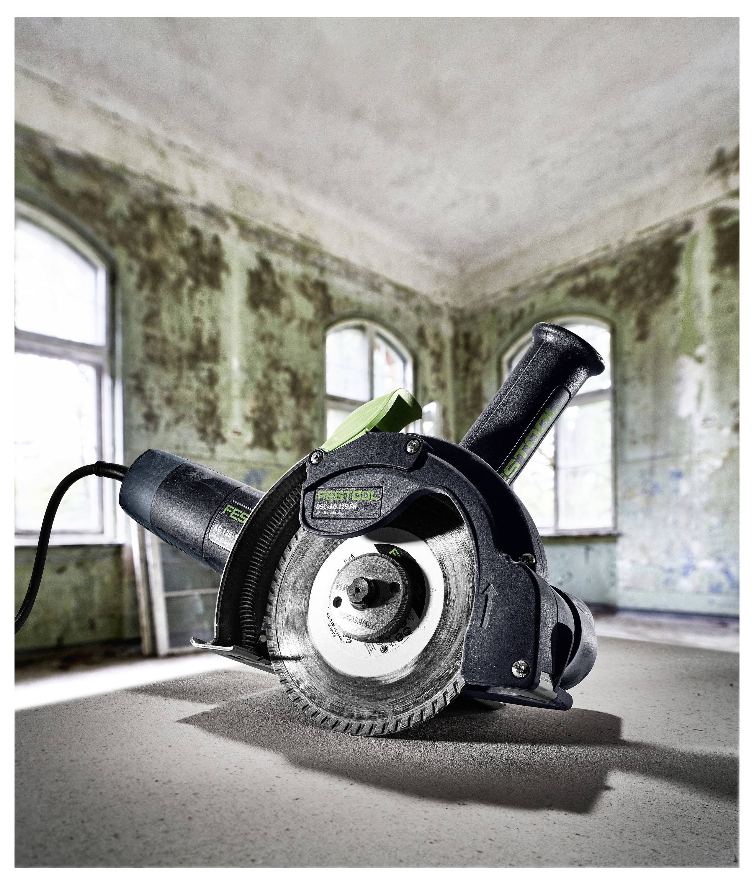Eine Kreissäge mit grünem Akzent steht auf einem sonnenbeschienenen, staubigen Boden in einem verfallenen Raum mit abblätternder Farbe und großen Fenstern.
