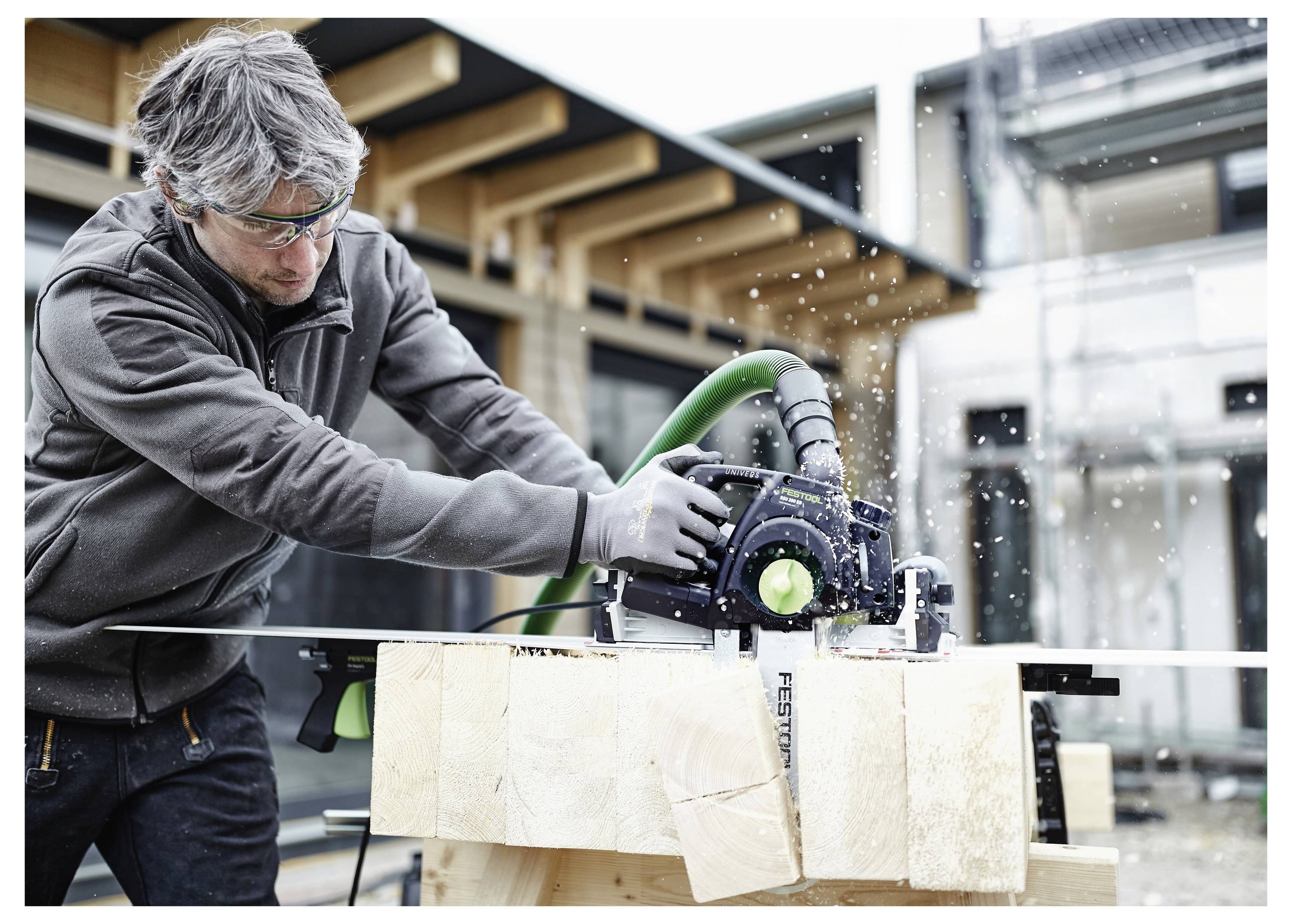Eine Person mit Schutzbrille bedient eine Kreissäge an einem Holzbalken, im Freien, wobei Sägespäne um sie herum verstreut werden.