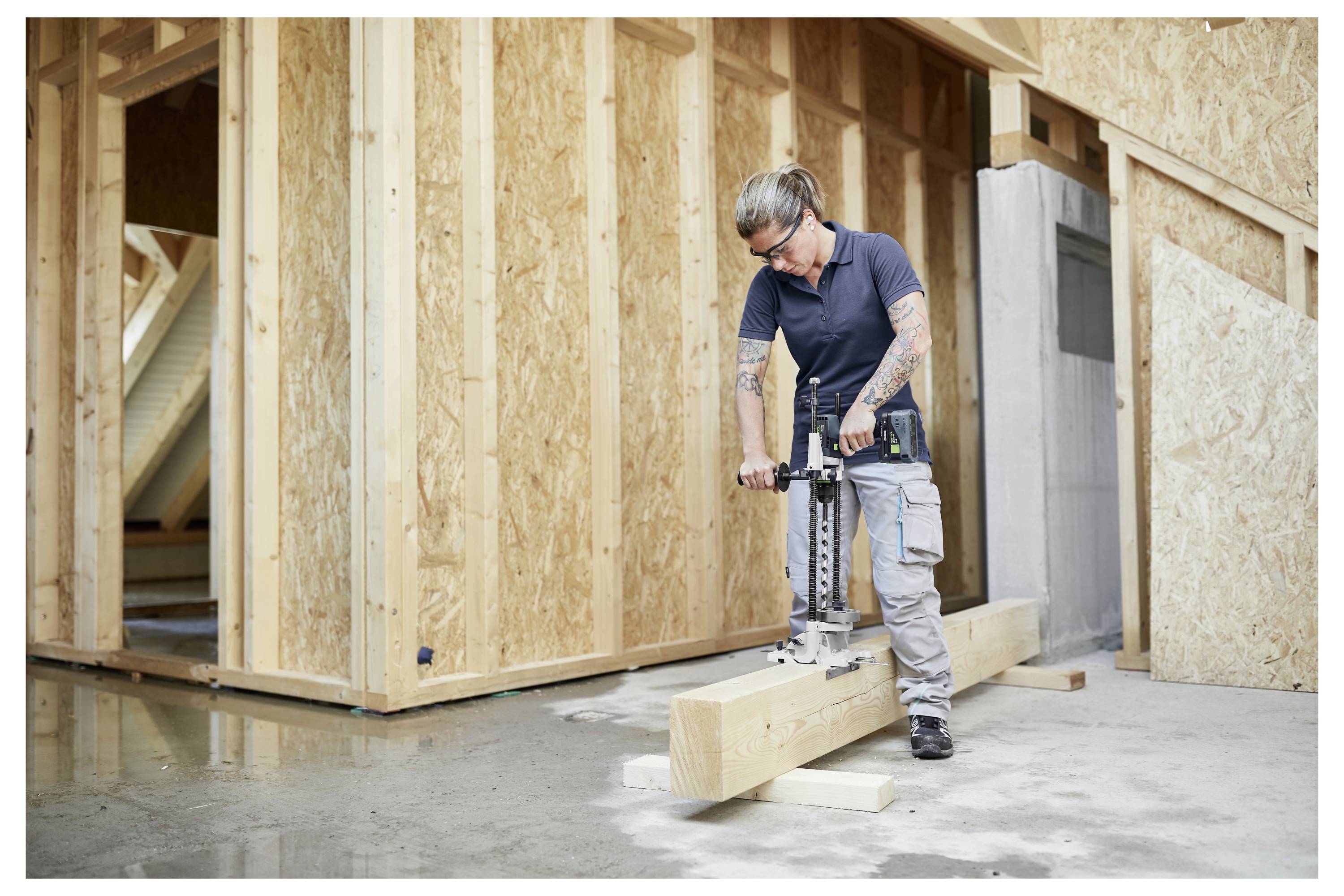 Eine Person arbeitet an einem Holzbalken in einem teilweise konstruierten Gebäude und verwendet ein Werkzeug, um präzise Anpassungen oder Schnitte vorzunehmen.
