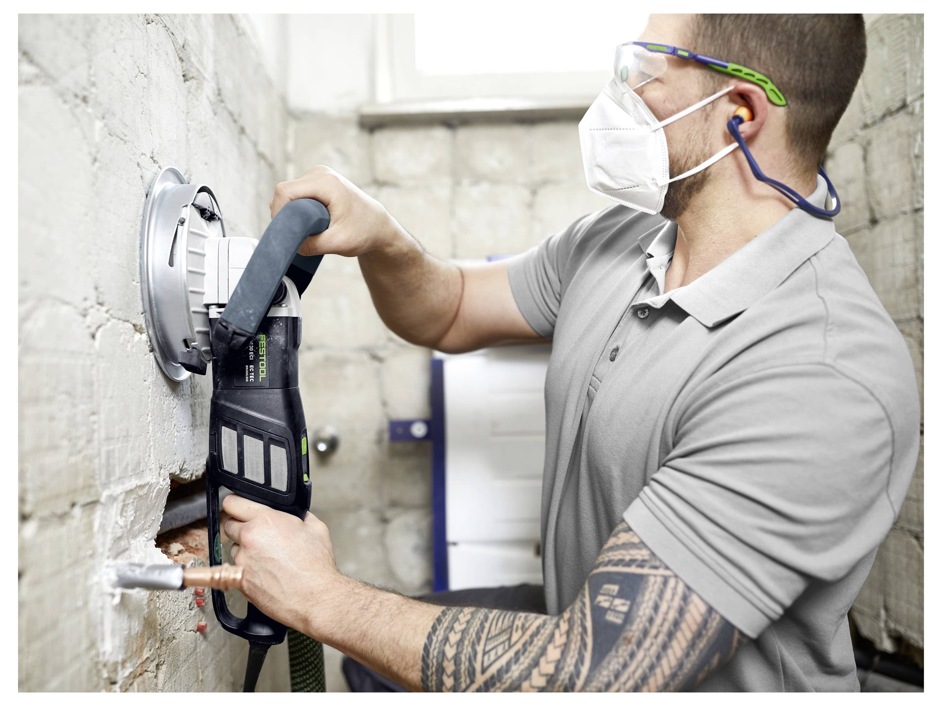 Eine Person in Schutzausrüstung nutzt ein Elektrowerkzeug, um in eine weiße Ziegelmauer zu schneiden und legt dabei Rohre frei.