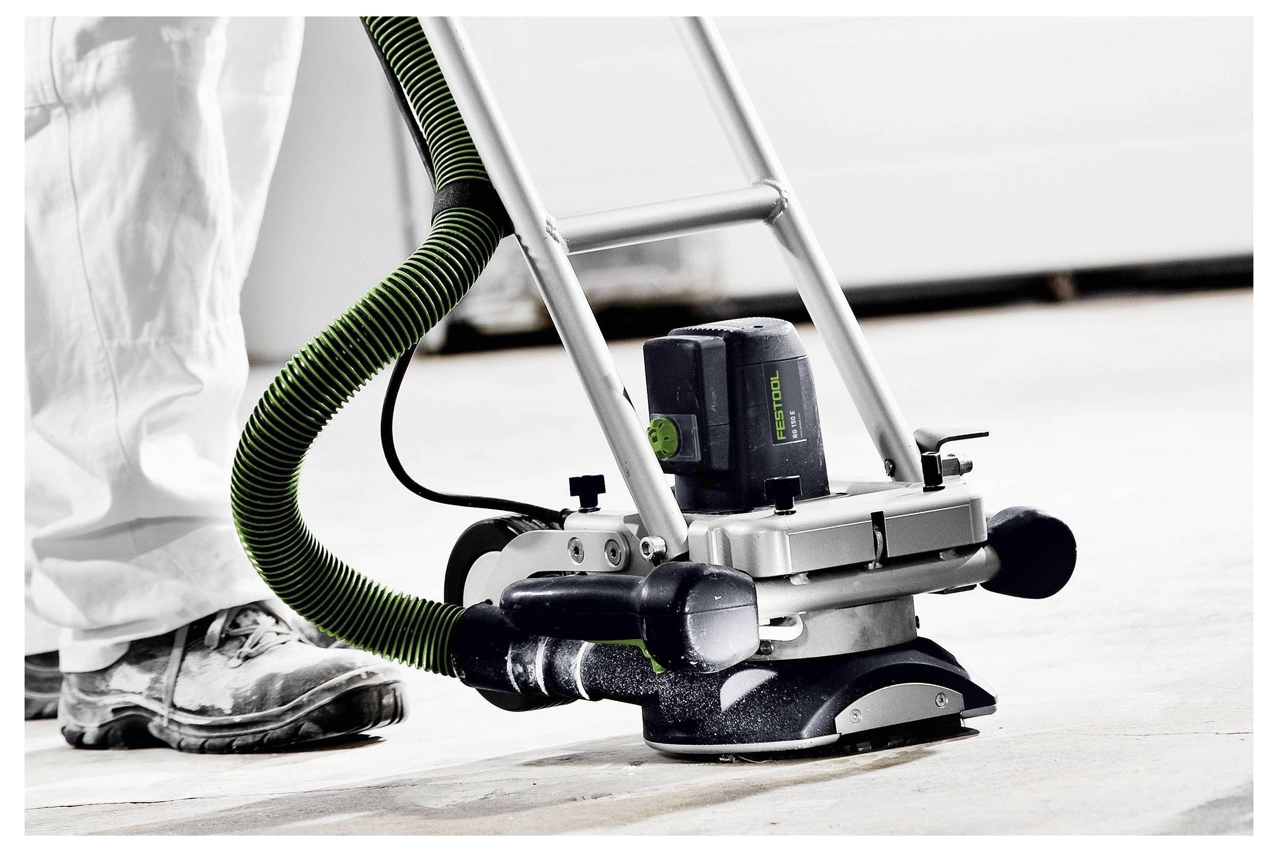 Eine Nahaufnahme einer Person, die eine Bodenschleifmaschine mit einem grünen Staubsaugerschlauch bedient und eine Betonoberfläche in Innenräumen glättet.