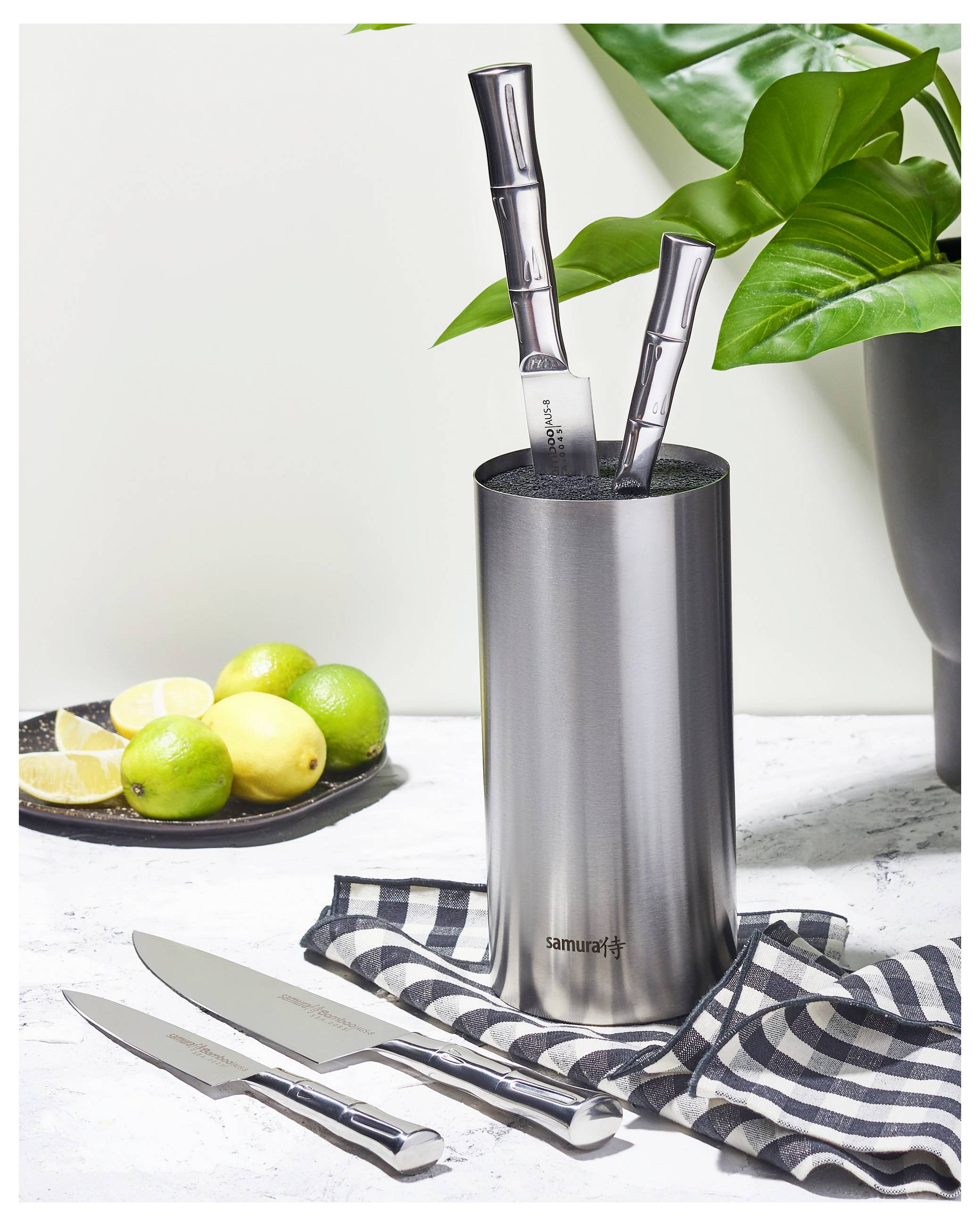 A set of stainless steel kitchen knives in a cylindrical holder on a marble surface with a striped cloth. A bowl with lemons and limes is nearby.