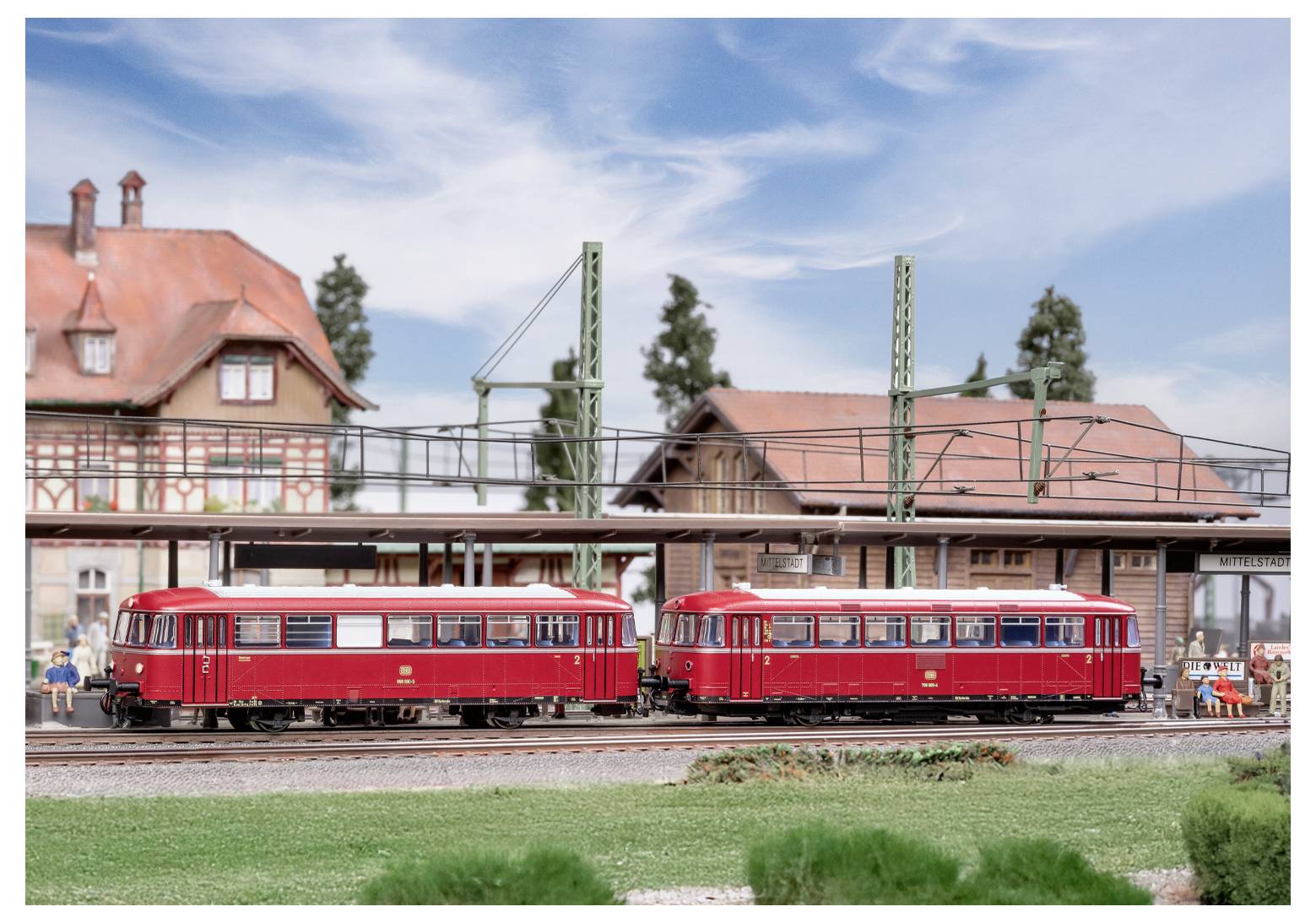 Zwei rote Modelleisenbahnen auf Gleisen in einer detaillierten Miniatur-Dorflandschaft mit Häusern und Bäumen unter einem klaren blauen Himmel.