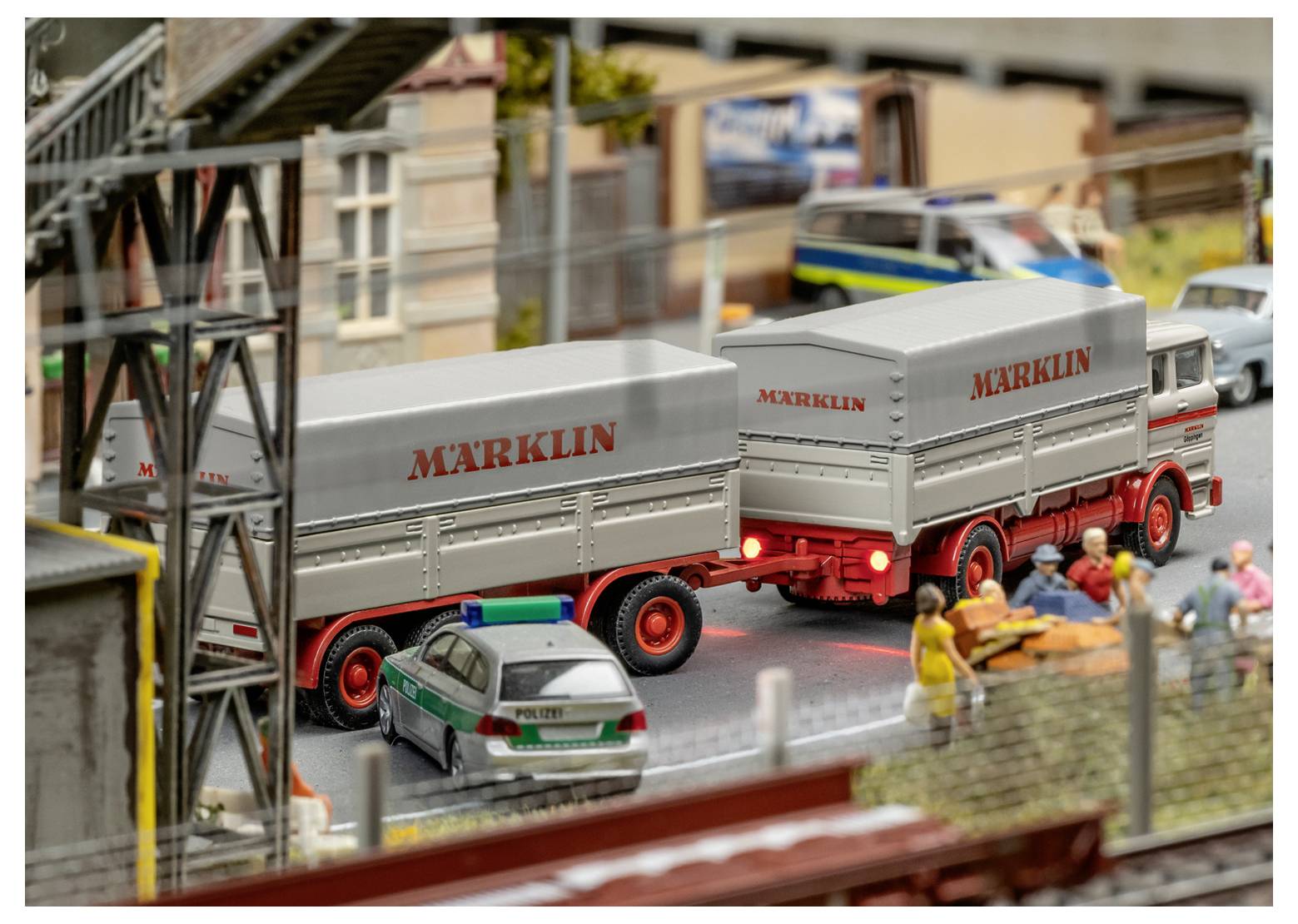 Eine Modellszene mit zwei Märklin-Lastkraftwagen auf einer Straße und einem Polizeiauto in der Nähe. Menschen versammeln sich am Straßenrand in einer städtischen Umgebung.