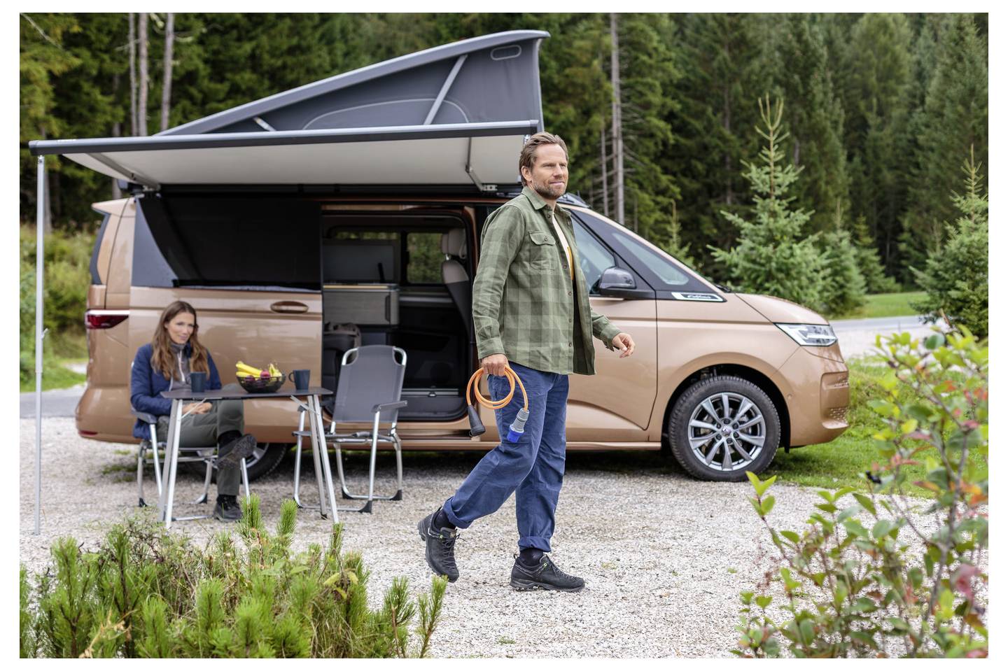 Ein Mann trägt ein orangefarbenes Kabel neben einem Wohnmobil mit Hubdach, während eine Frau an einem kleinen Tisch außerhalb auf einem Waldcampingplatz sitzt.