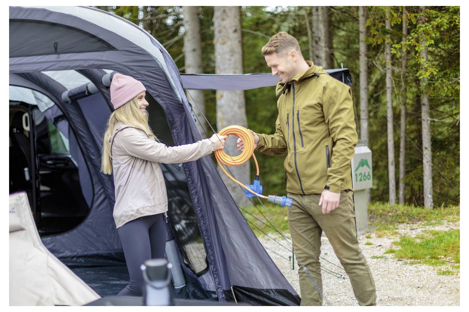 Mann und Frau tauschen an einem Campingplatz ein orangefarbenes Verlängerungskabel neben einem offenen Zelt in einem Waldgebiet aus.