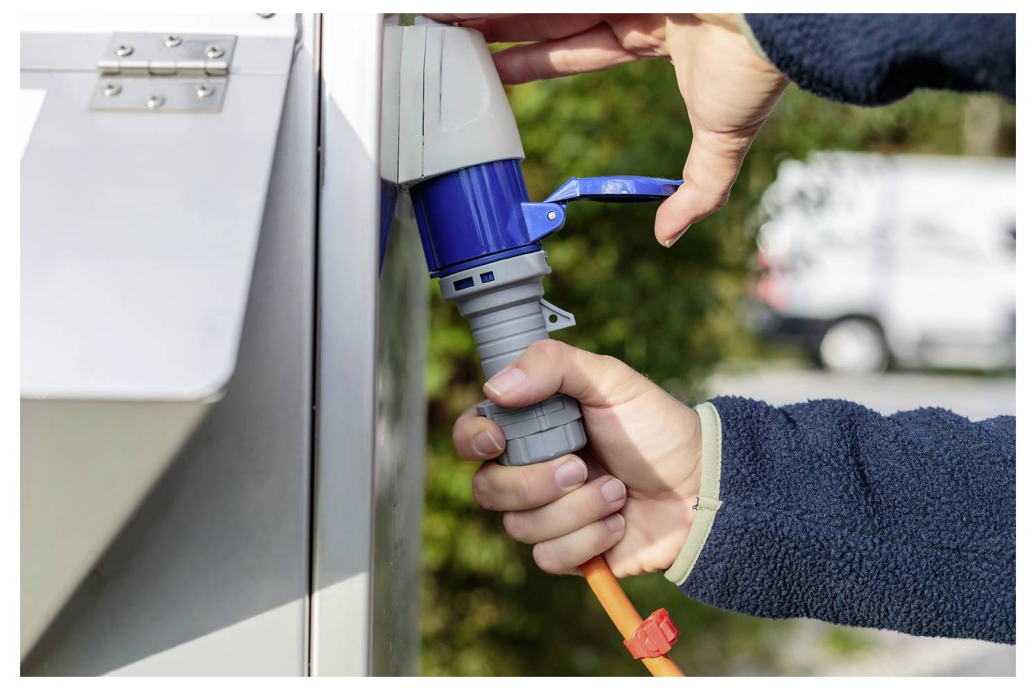 Eine Person steckt ein orangefarbenes Stromkabel in eine Außensteckdose, was darauf hindeutet, dass etwas im Freien eingerichtet oder mit Strom versorgt wird.