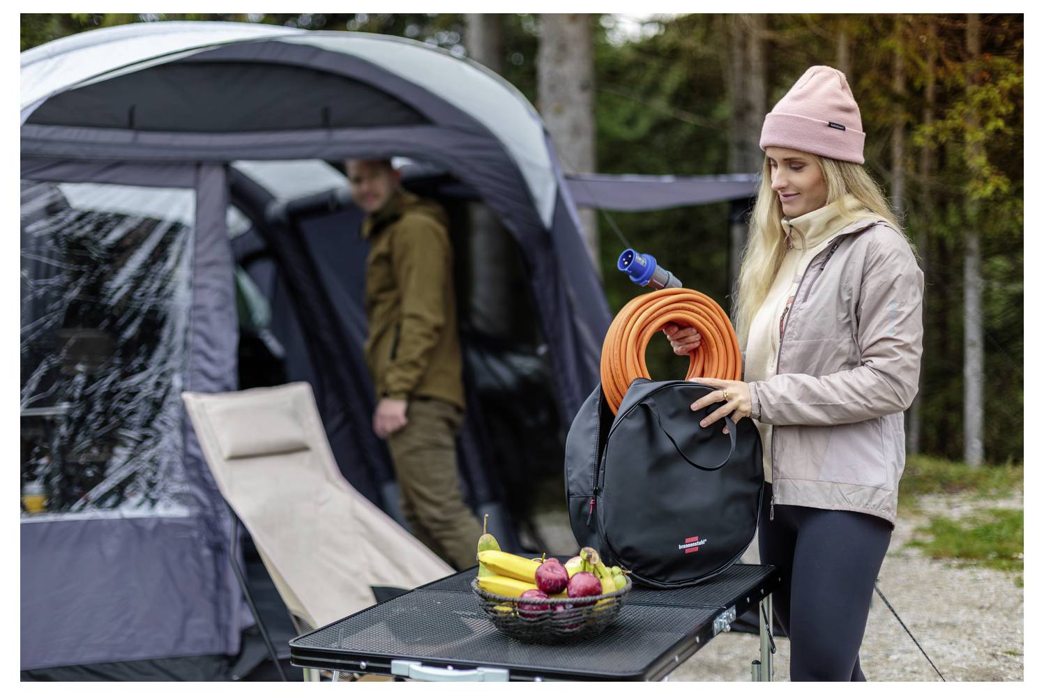 Frau packt Campingausrüstung neben einem Zelt und hält einen Rucksack mit einem aufgerollten orangefarbenen Schlauch; Picknicktisch mit Obstkorb im Vordergrund.