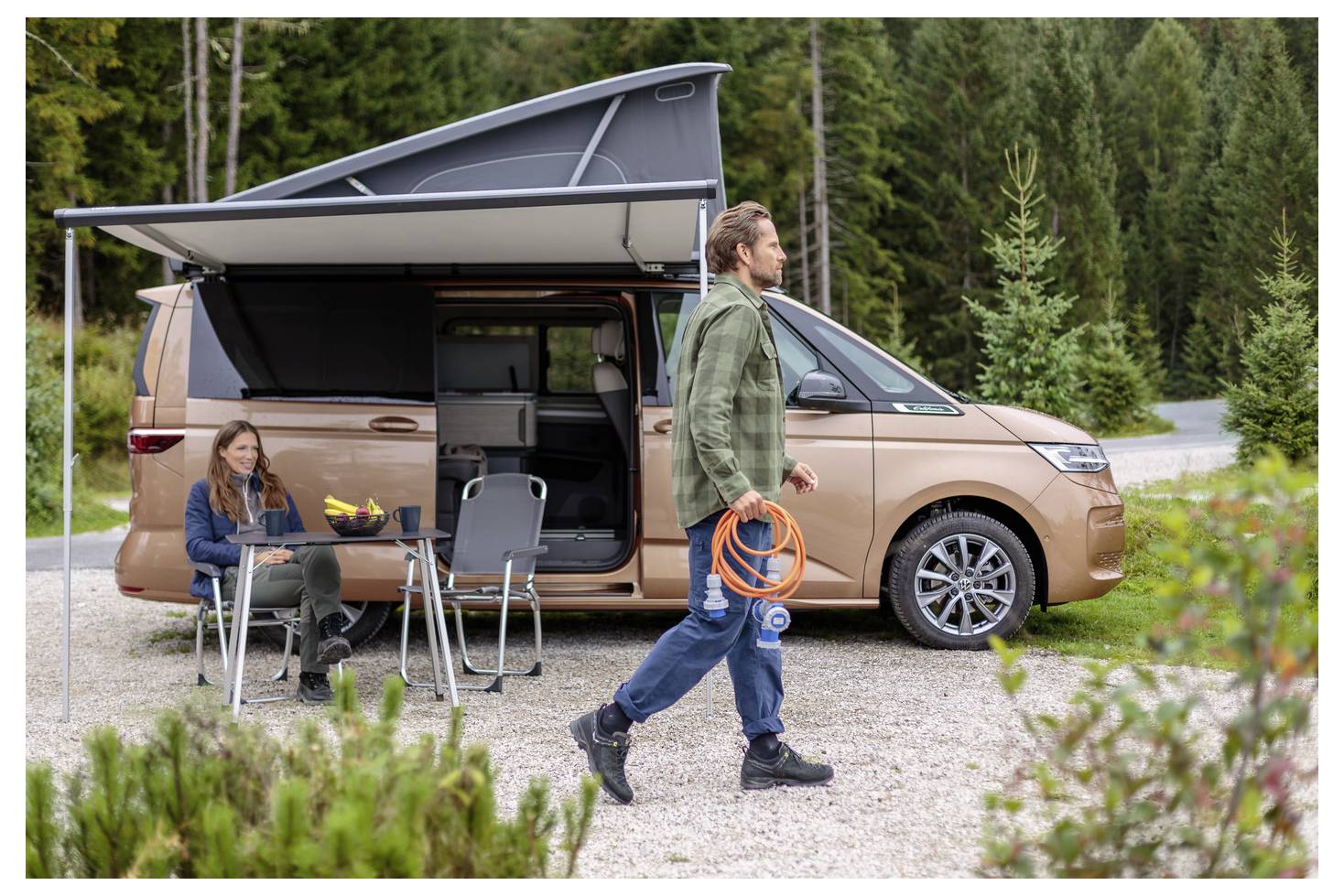 Ein Paar genießt das Camping neben einem beigen Wohnmobil in einem bewaldeten Gebiet. Eine Person sitzt an einem Tisch, während die andere ein Verlängerungskabel trägt.