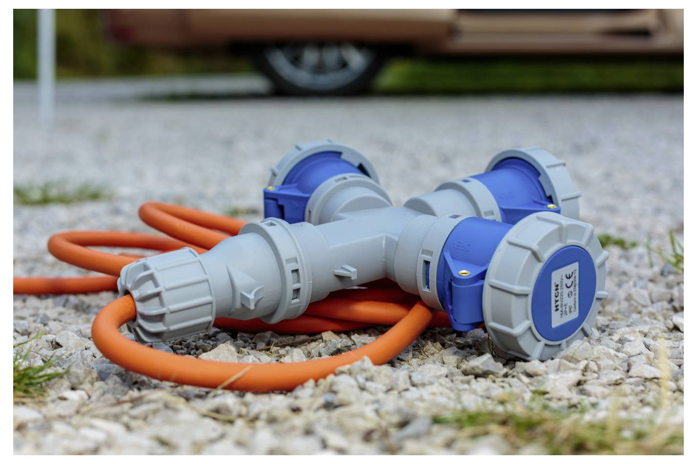 Ein orangefarbenes elektrisches Verlängerungskabel mit mehreren blauen und grauen Steckdosen liegt aufgerollt auf einer Kiesfläche in der Nähe eines Fahrzeugs.