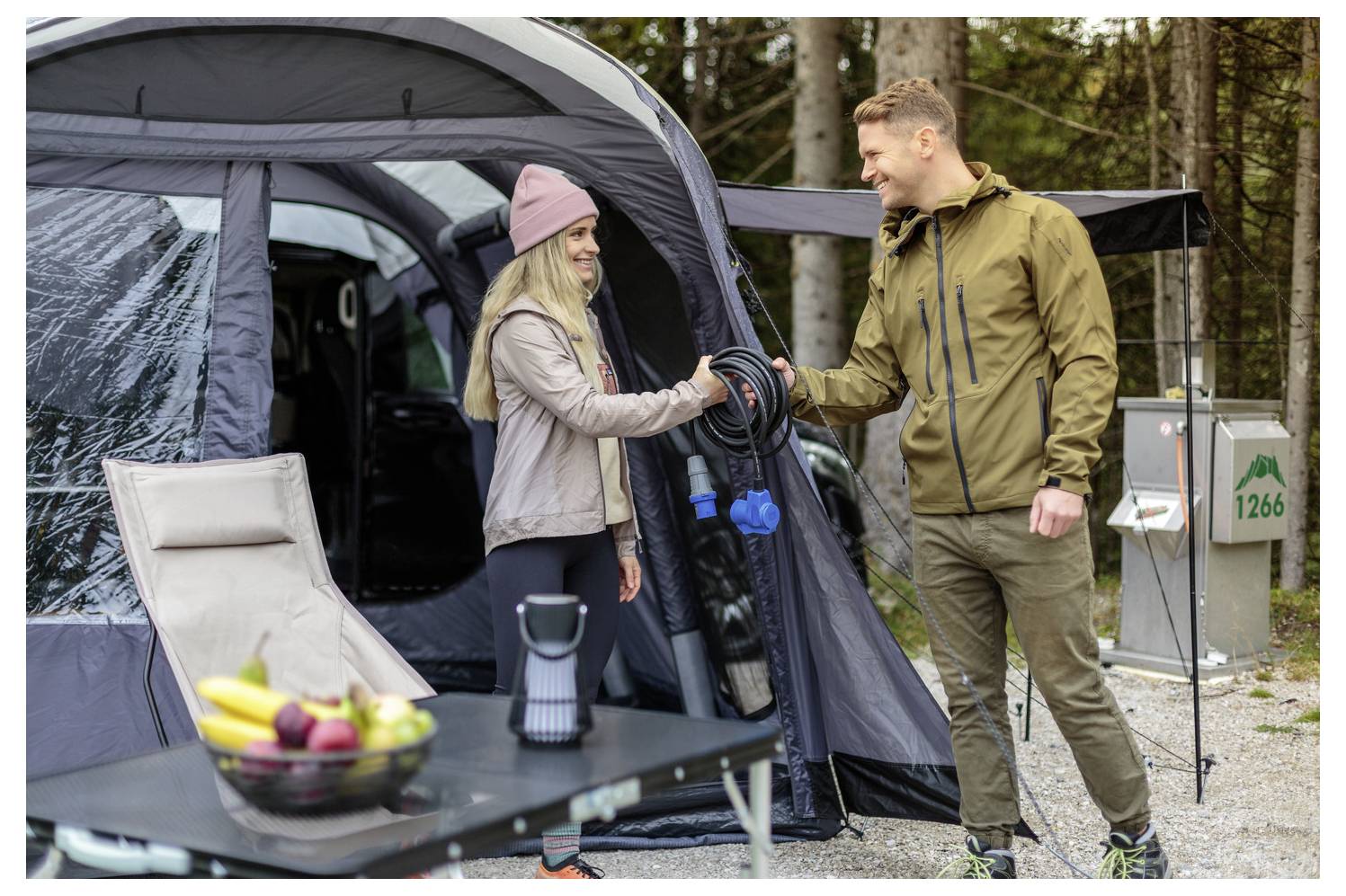 Ein Mann und eine Frau lächeln und schütteln sich die Hände außerhalb eines Zeltes in einem Campingplatz, mit sichtbarem Campingequipment um sie herum.