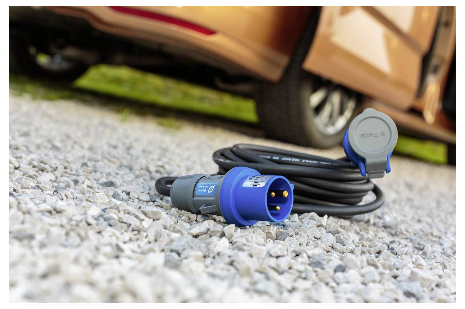 Ein blauer Elektrostecker mit einem langen schwarzen Kabel liegt auf Kieselsteinen in der Nähe des Rades eines geparkten braunen Fahrzeugs.