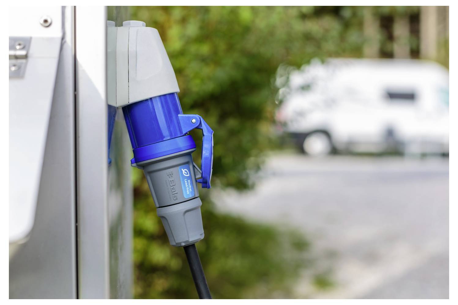 Ein blauer Elektro-Ladestecker, der an eine Steckdose an einem Gebäude angeschlossen ist, in einer grünen Außenumgebung mit einem unscharfen Fahrzeug im Hintergrund.