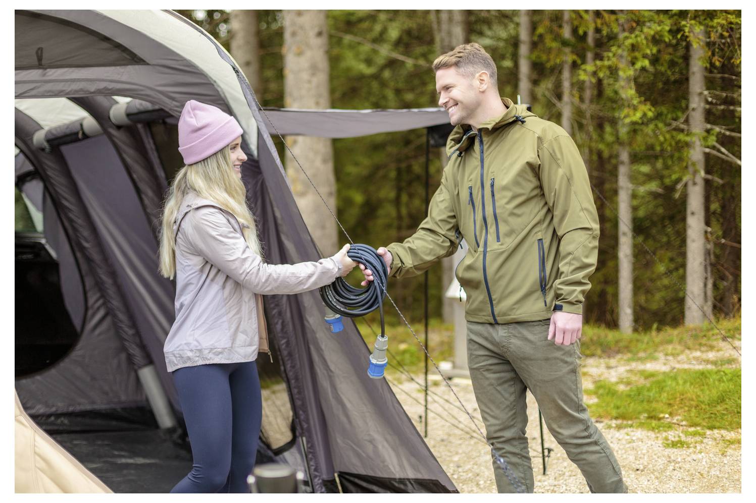 Eine Frau und ein Mann neben einem Zelt tauschen ein aufgerolltes Kabel aus; beide lächeln und tragen Outdoor-Jacken. Im Hintergrund sind Bäume und Gras.