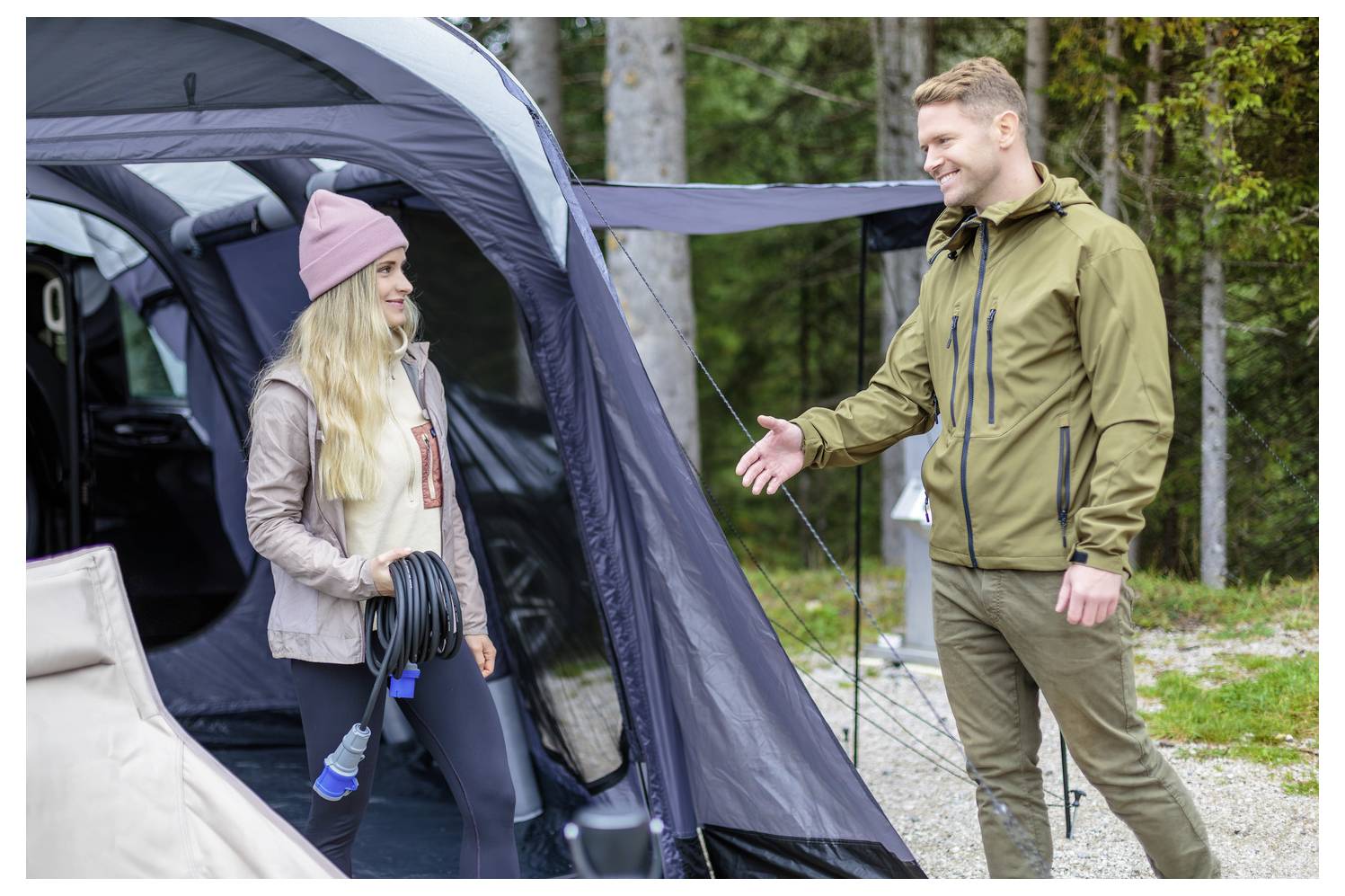 Eine Person steht außerhalb eines Zelts, hält ein aufgerolltes Kabel in der Hand und lächelt eine andere Person an, die ihre Hand zum Gruß ausstreckt.