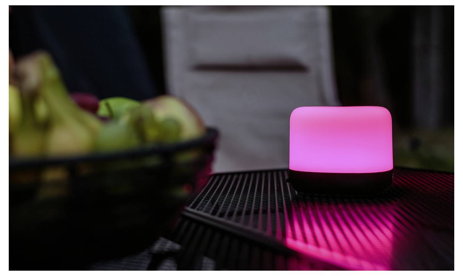 Eine leuchtend rosa Lampe auf einem Tisch neben einer Obstschale in einer schwach beleuchteten Umgebung, die eine gemütliche oder intime Atmosphäre vermittelt.