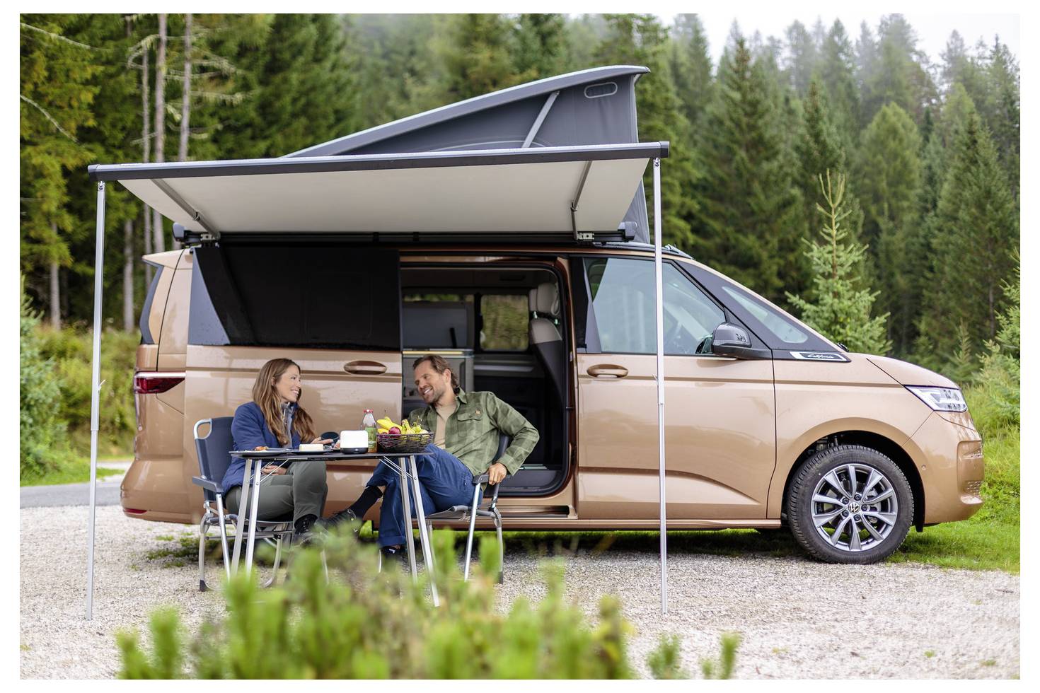 Ein brauner Wohnwagen mit erhöhtem Dach und Markise ist im Wald geparkt. Zwei Personen sitzen draußen an einem Tisch und genießen Getränke.