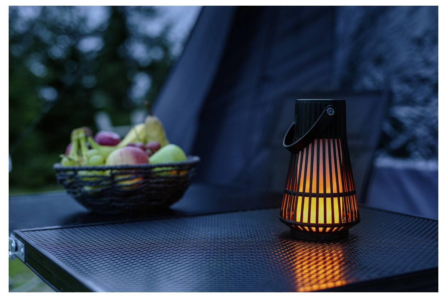 Eine leuchtende Laterne auf einem Außentisch neben einem Korb mit Äpfeln und Birnen in der Abenddämmerung, die eine gemütliche und einladende Atmosphäre schafft.